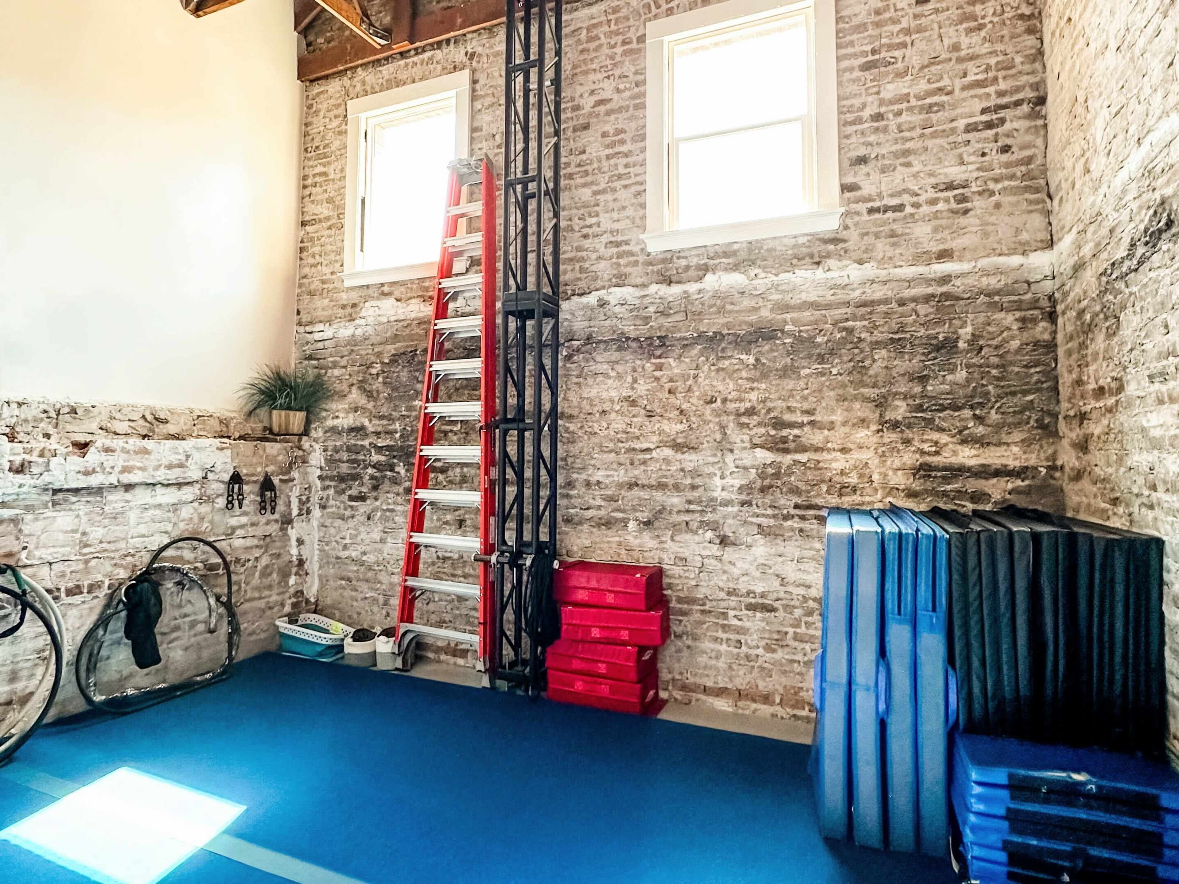 A workout space with a blue mat, a red ladder against a brick wall, and stacked exercise mats on the floor.