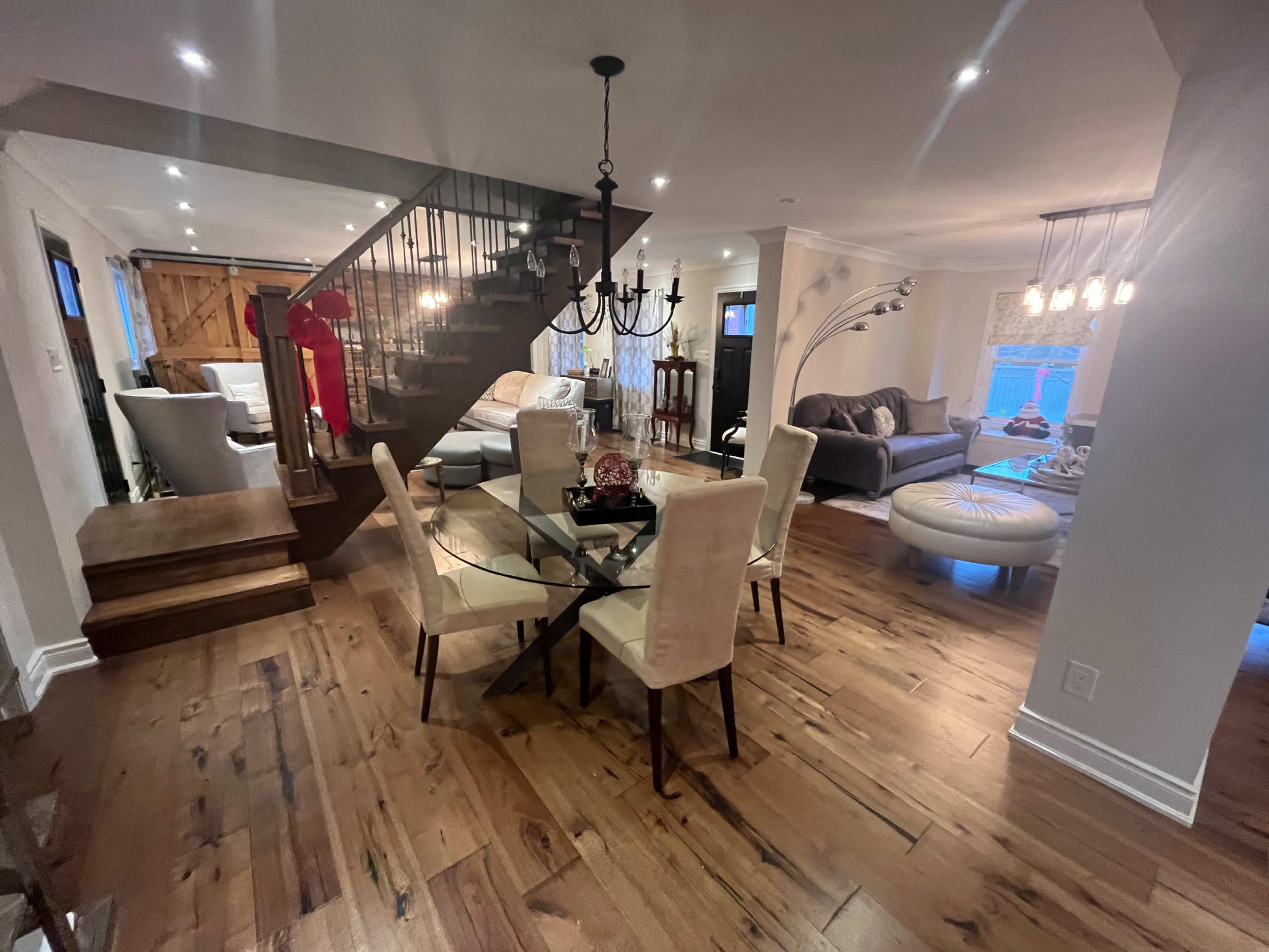 The image depicts an interior space featuring a dining area with a glass table surrounded by beige chairs, a staircase leading to an upper level, and a living area with various couches and decorative lighting.
