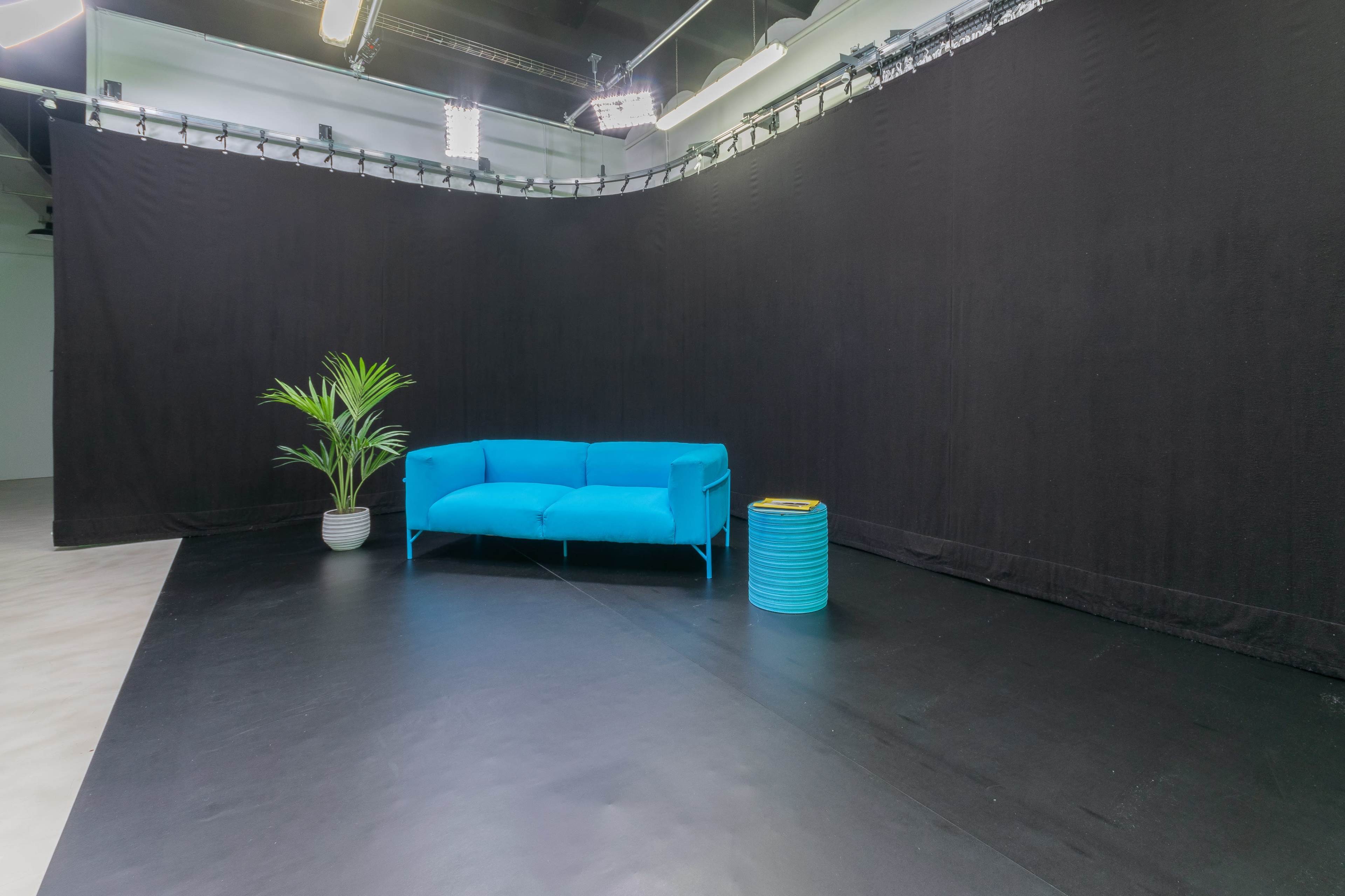 A bright blue sofa and a teal accent table are positioned in front of a black backdrop in a spacious studio setting.