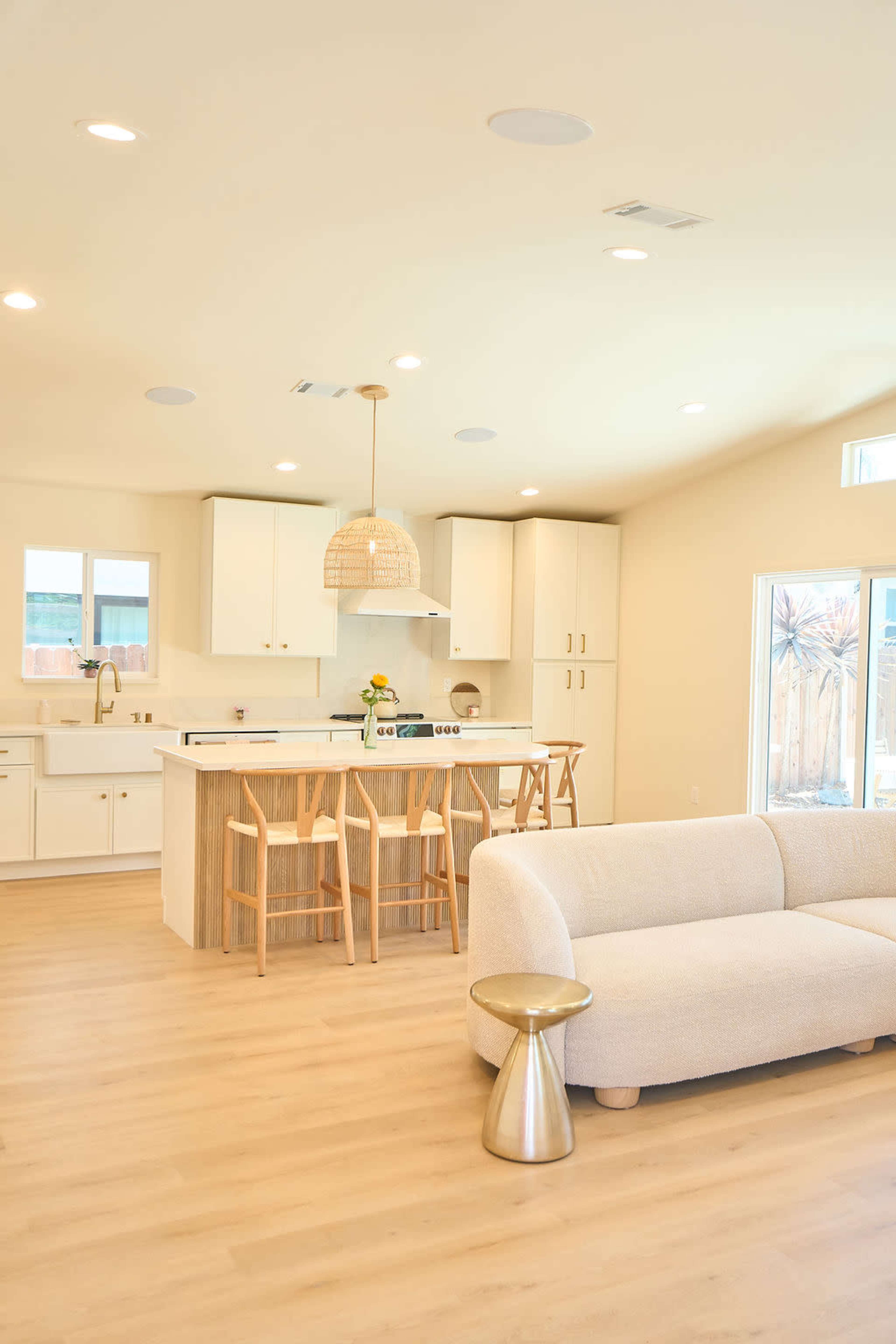 The image shows a bright and modern kitchen and living area with light wood flooring, white cabinetry, and a cozy sofa.