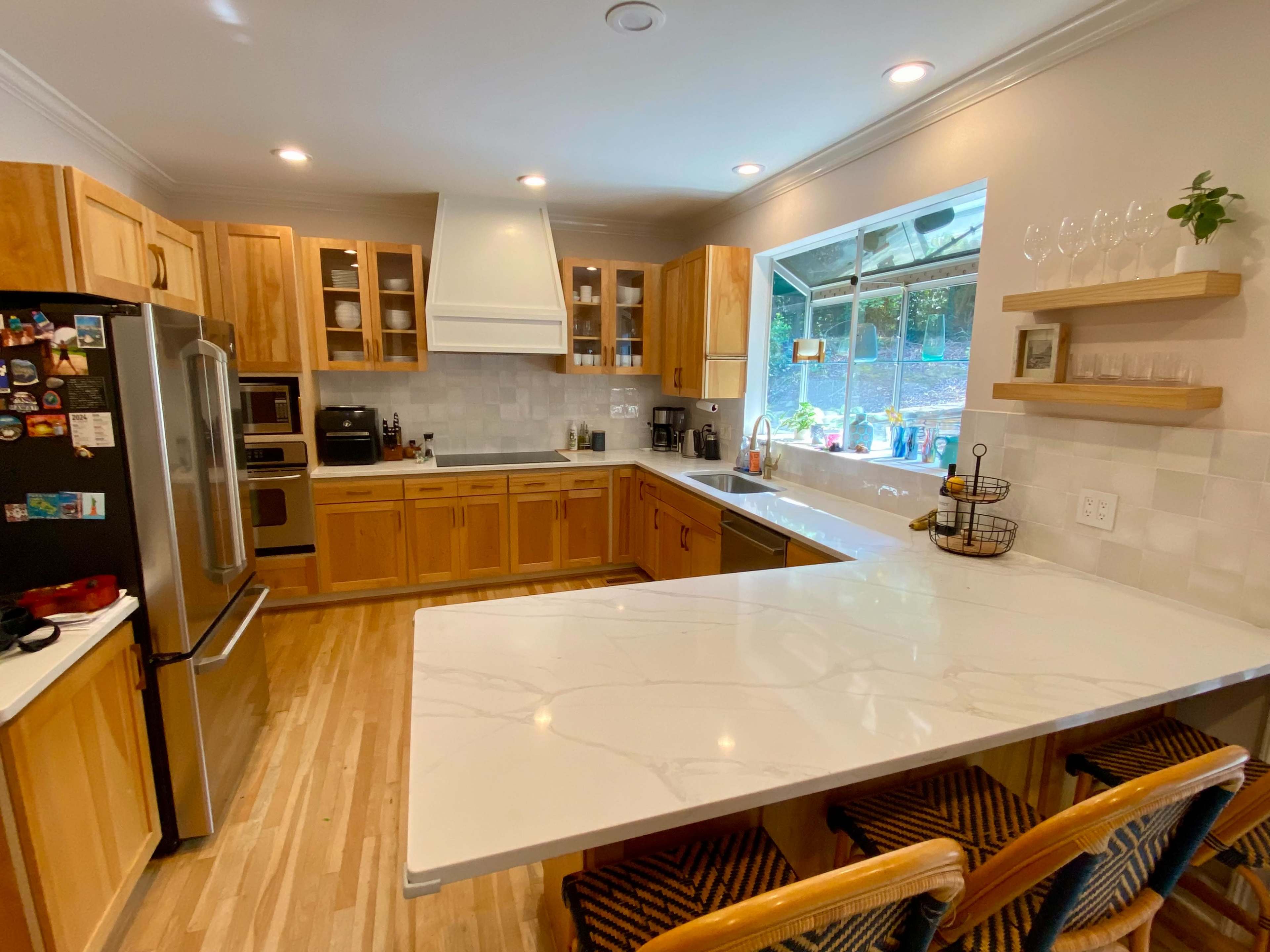 The image depicts a modern kitchen with wooden cabinets, stainless steel appliances, and a large white countertop with three wooden bar stools.