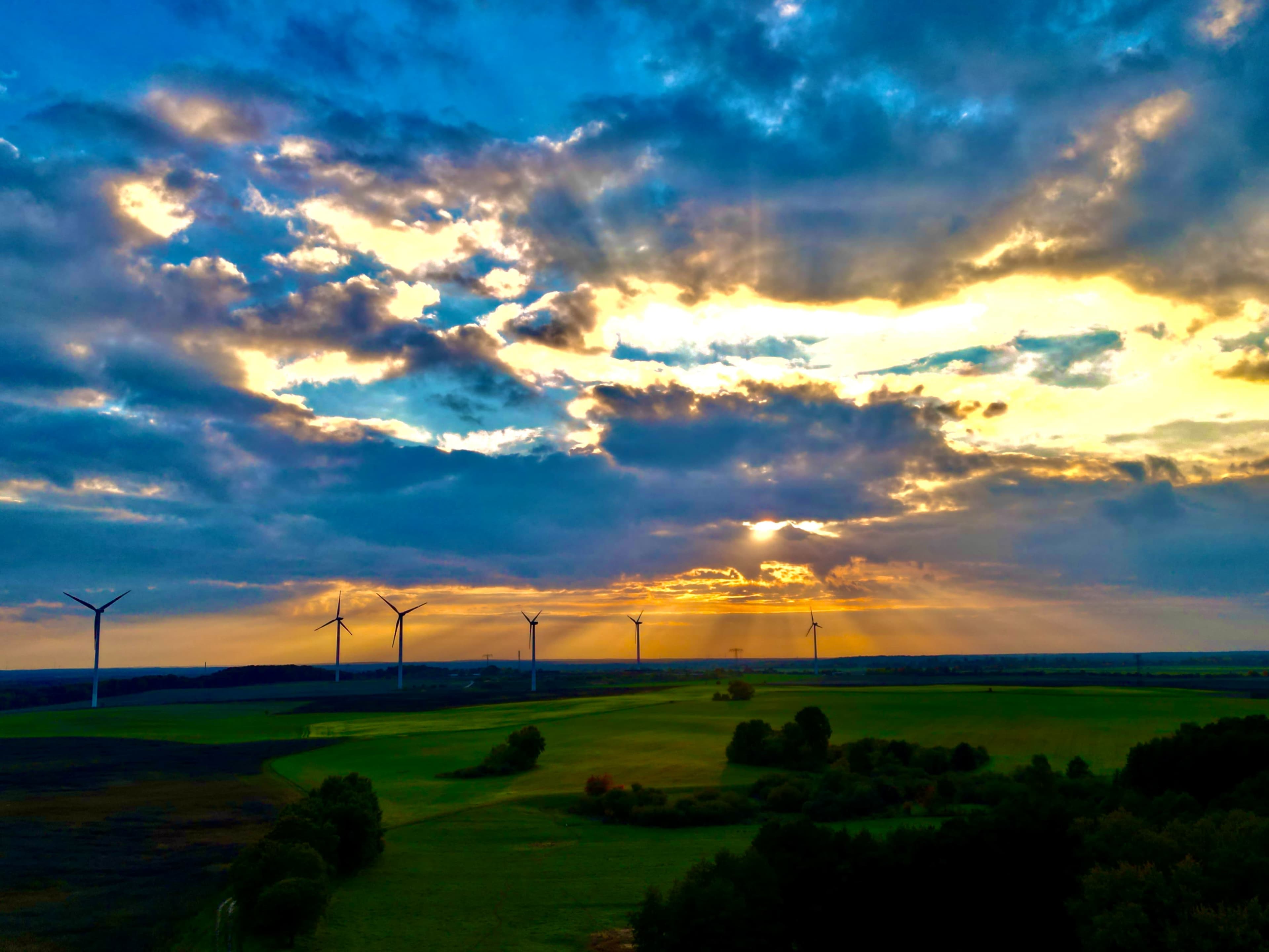 The image shows a landscape with wind turbines in the background under a sky filled with dramatic clouds and rays of sunlight breaking through.