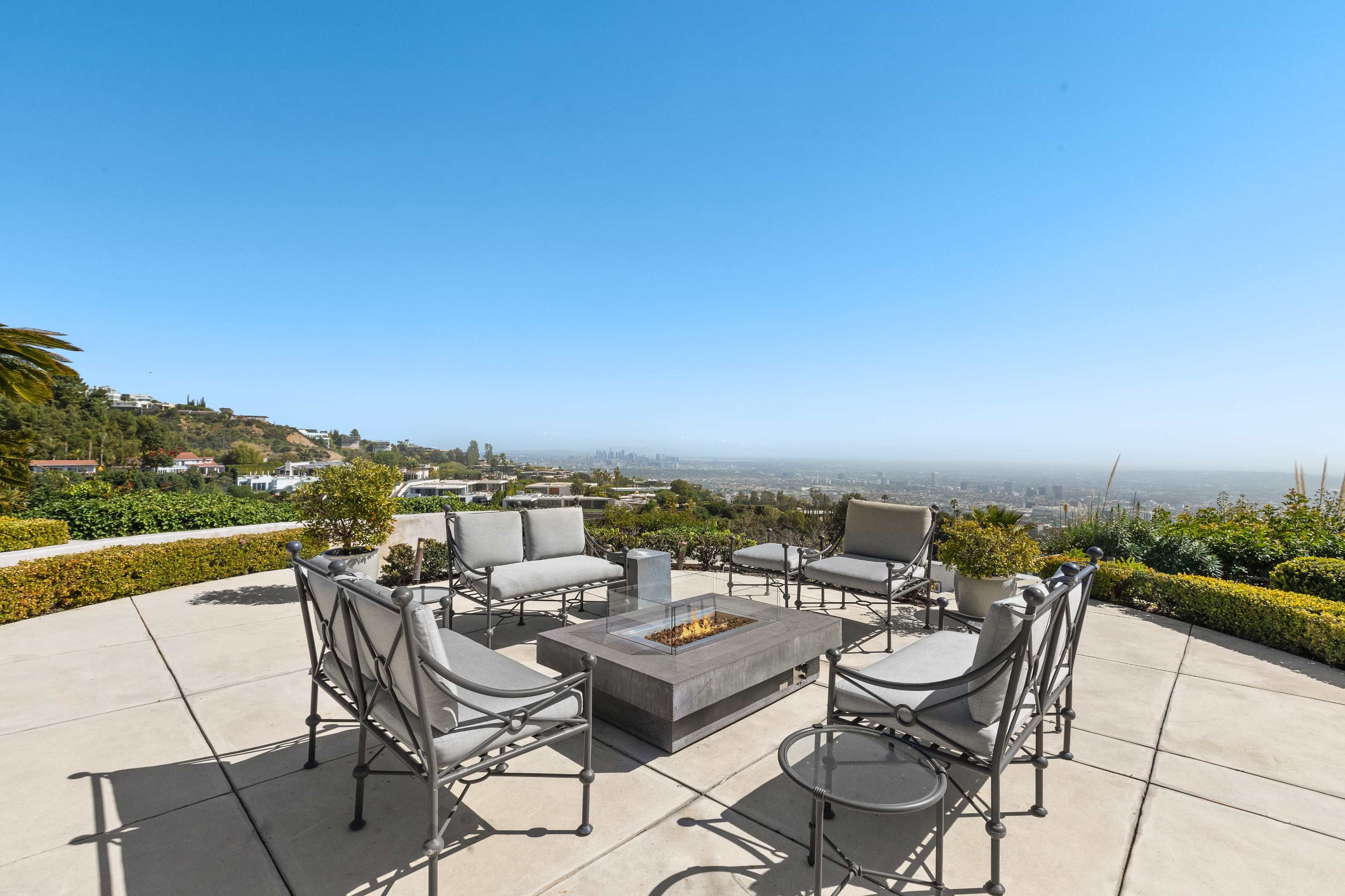 A patio features a central fire pit surrounded by several gray chairs, offering a panoramic view of the city below.