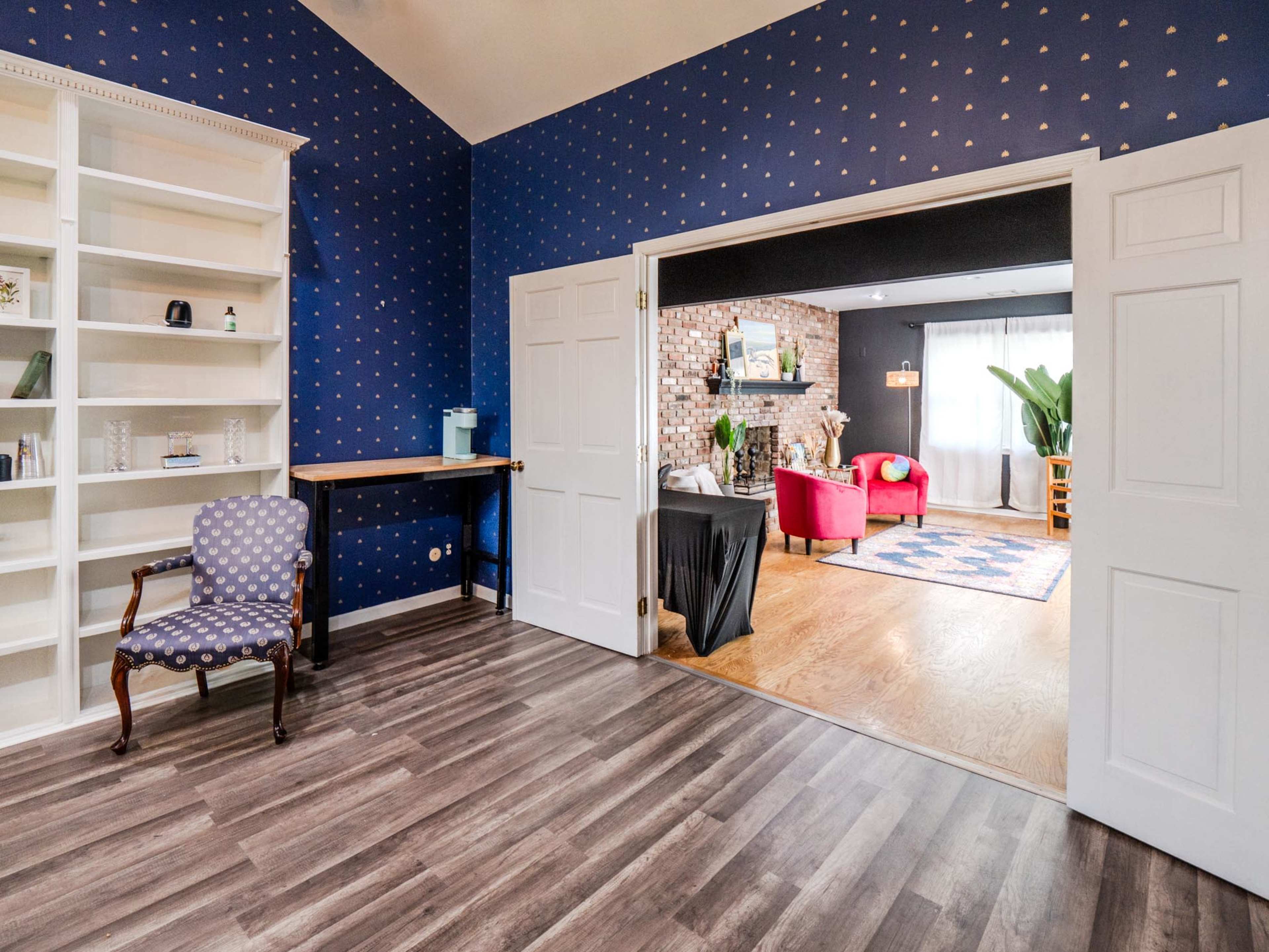 A room with dark blue walls decorated with gold polka dots features a bookshelf, a chair, and an open doorway leading to a brightly lit space with red furniture and a patterned rug.