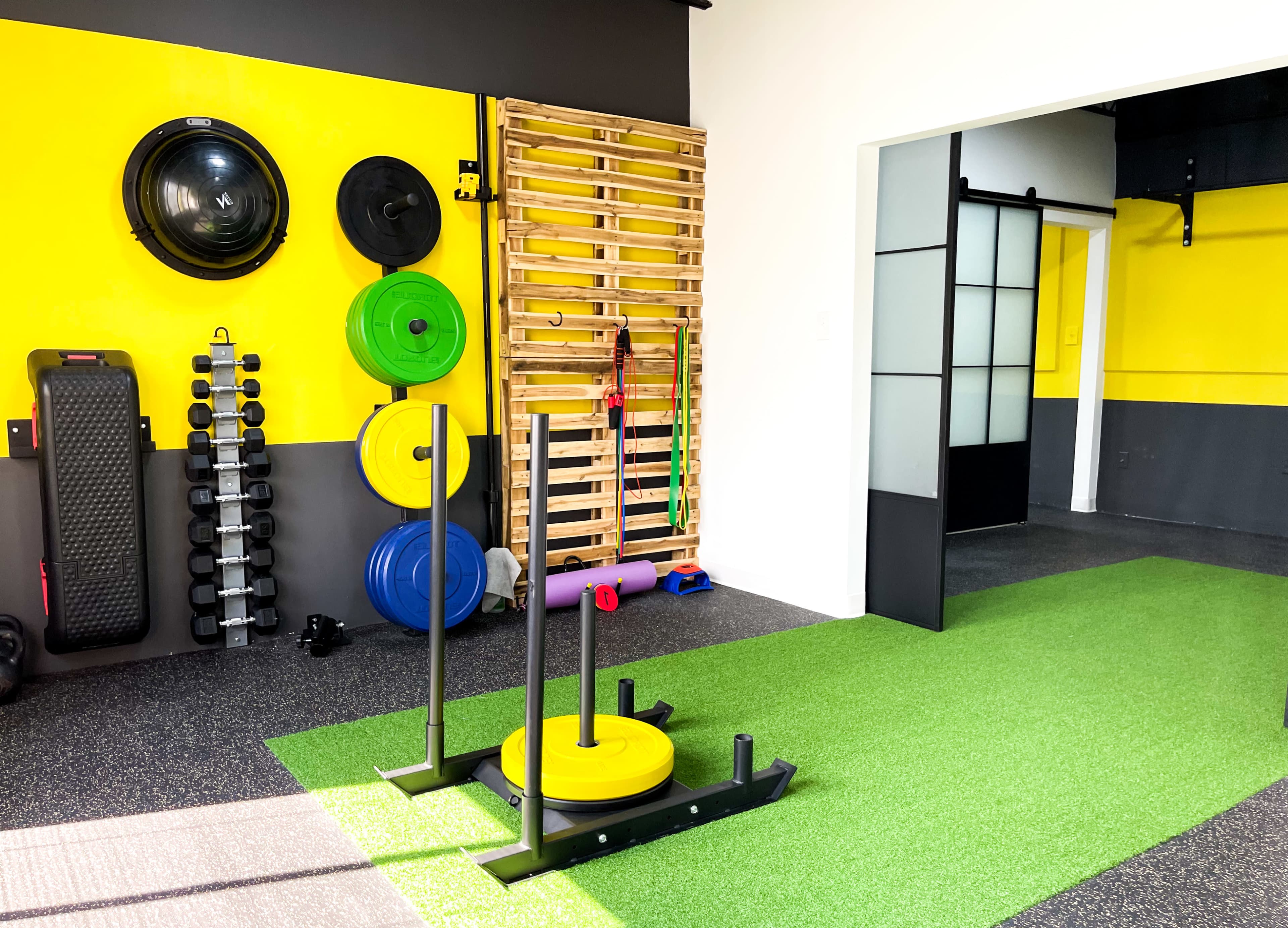 A fitness space featuring a sled for weighted exercises, various colored weights, a wall-mounted pegboard, and green turf flooring.