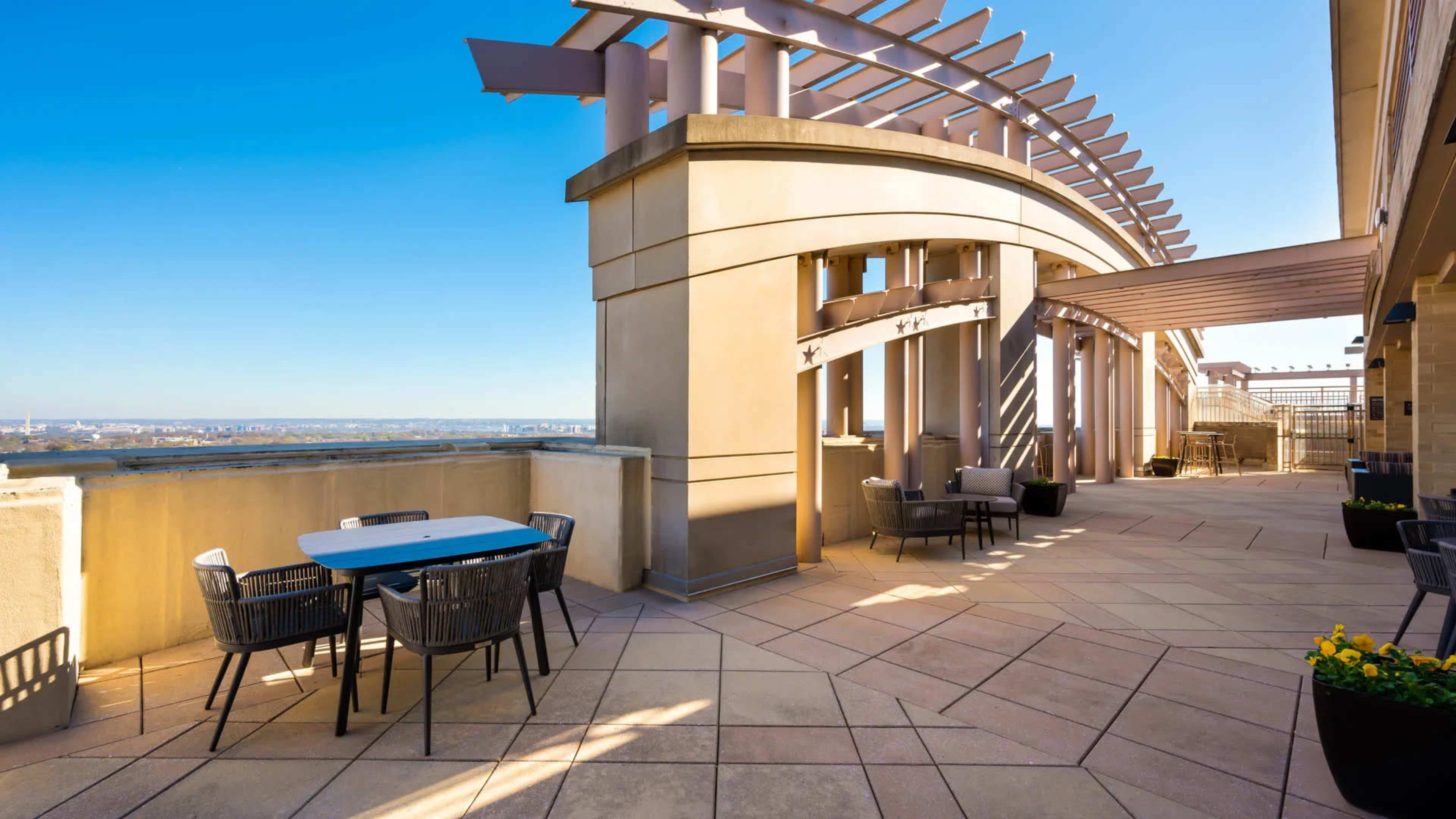 A spacious outdoor terrace area with a table and chairs, surrounded by architectural features and a clear blue sky.