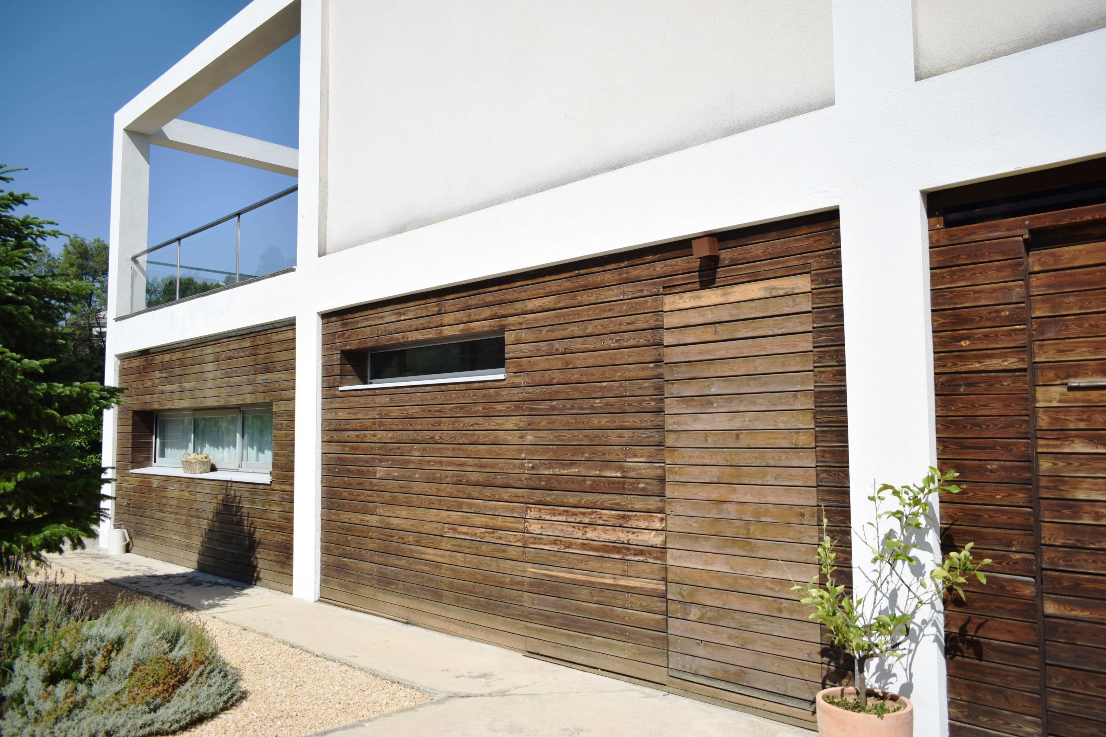 The image shows a modern house with a white exterior and wooden garage doors, surrounded by a landscaped area with gravel and greenery.