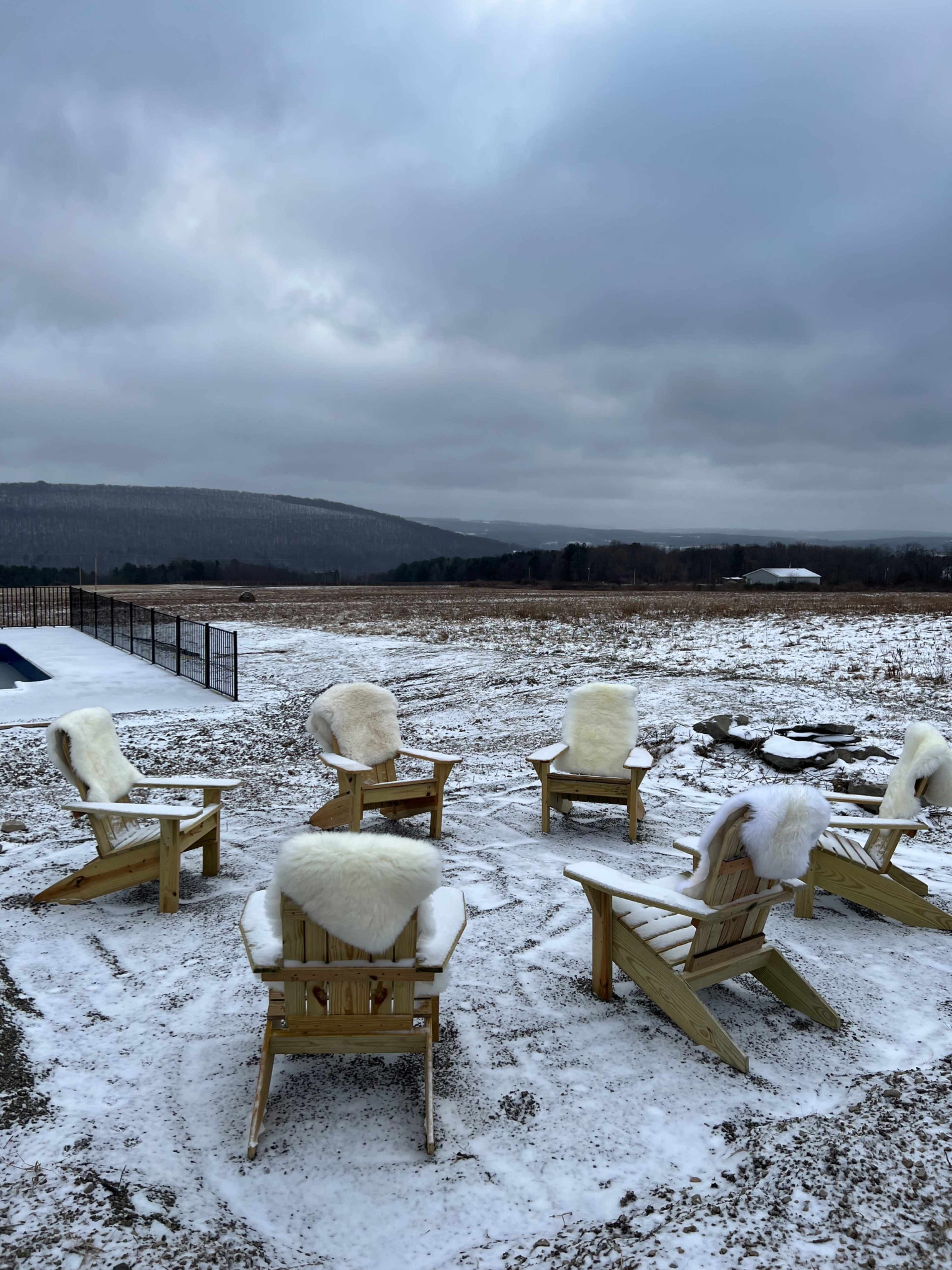 Six wooden adirondack chairs with fur cushions are arranged in a circle on a snowy landscape, with mountains in the background under a cloudy sky.
