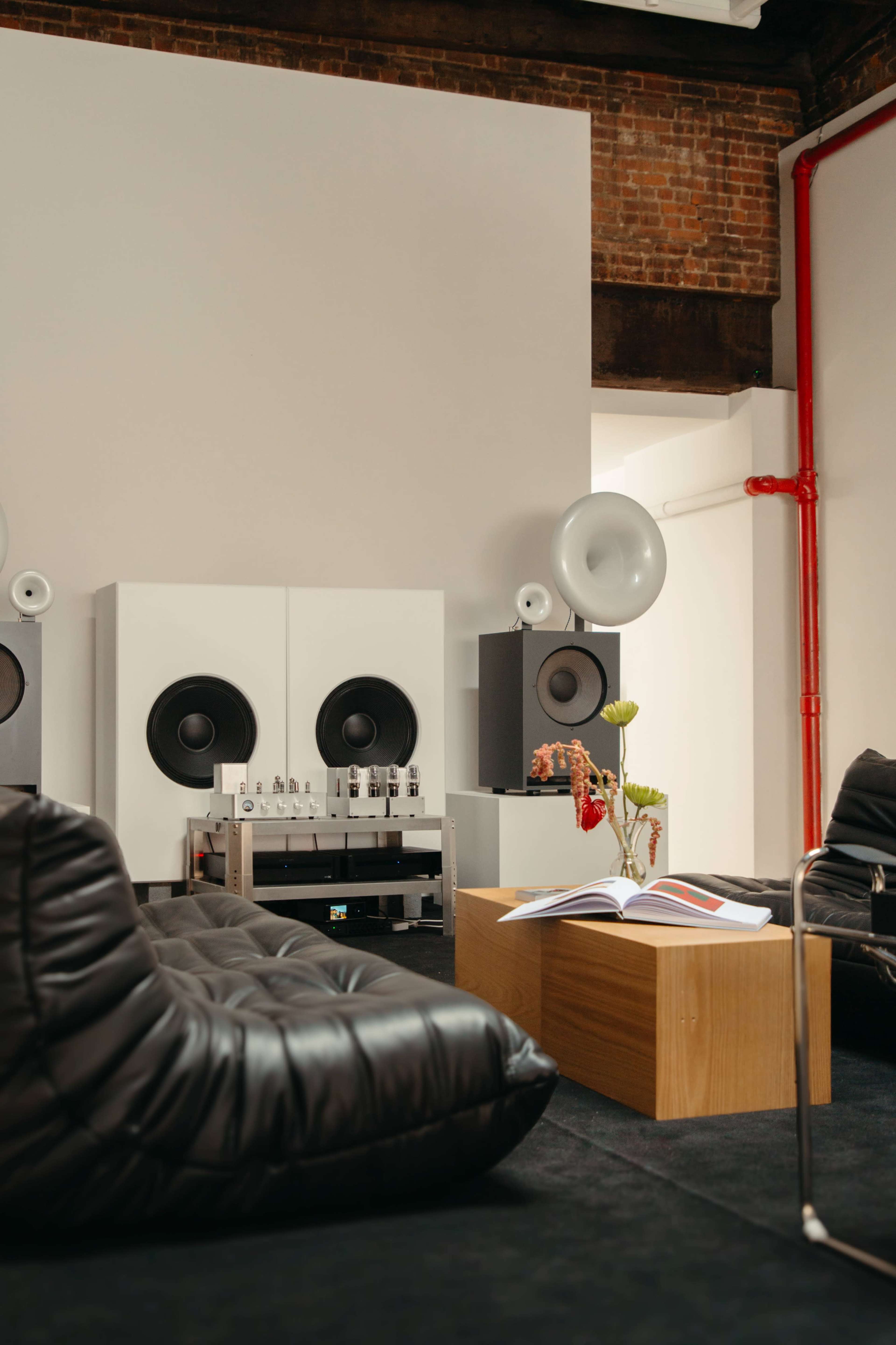 A minimalist lounge area features black beanbag chairs, a wooden coffee table with a book, and large audio speakers against a white wall in a brick-walled room.