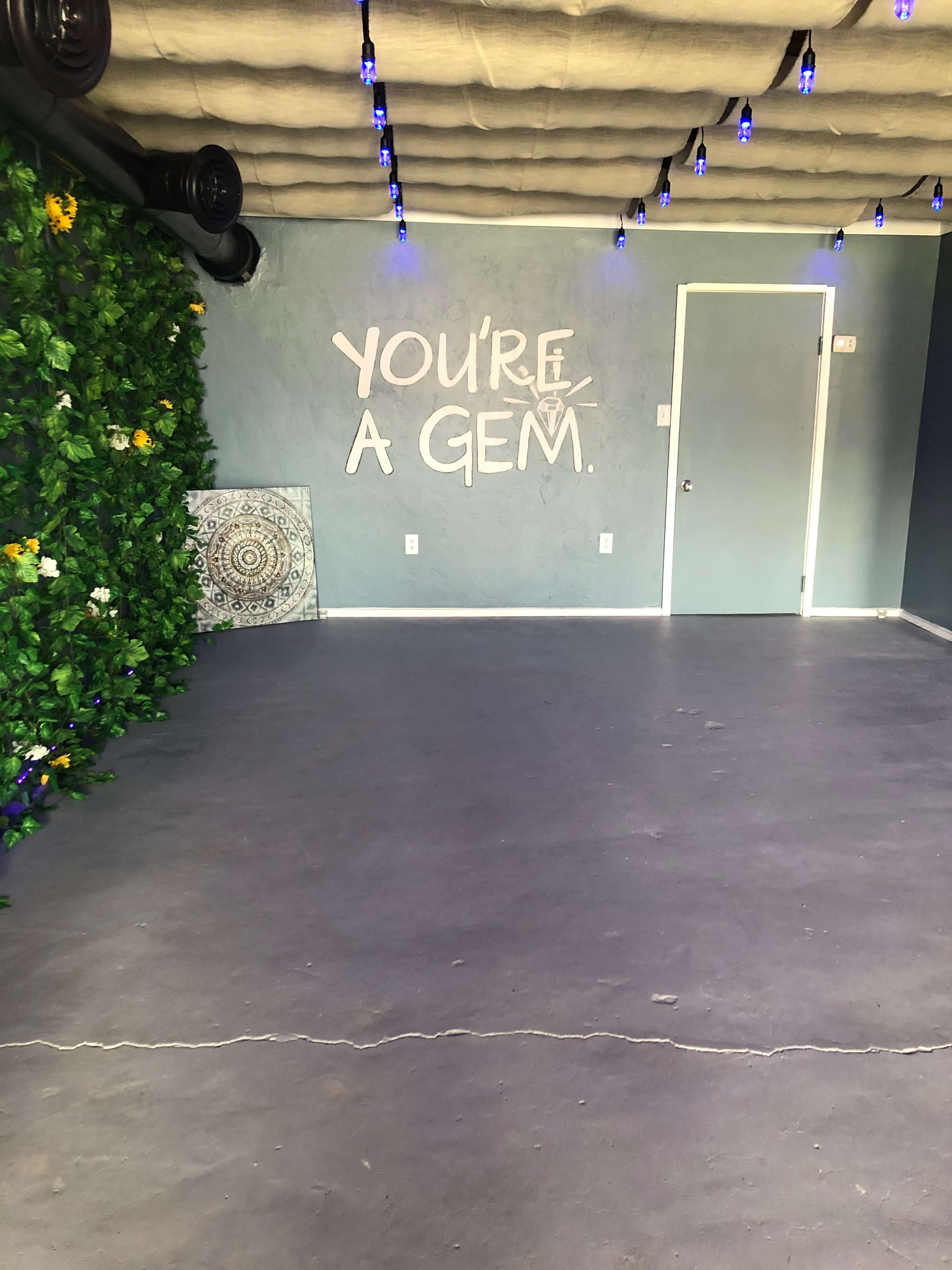 An empty room features a blue wall with the text "YOU'RE A GEM," decorative greenery on one side, and a partially visible mandala design on the floor.