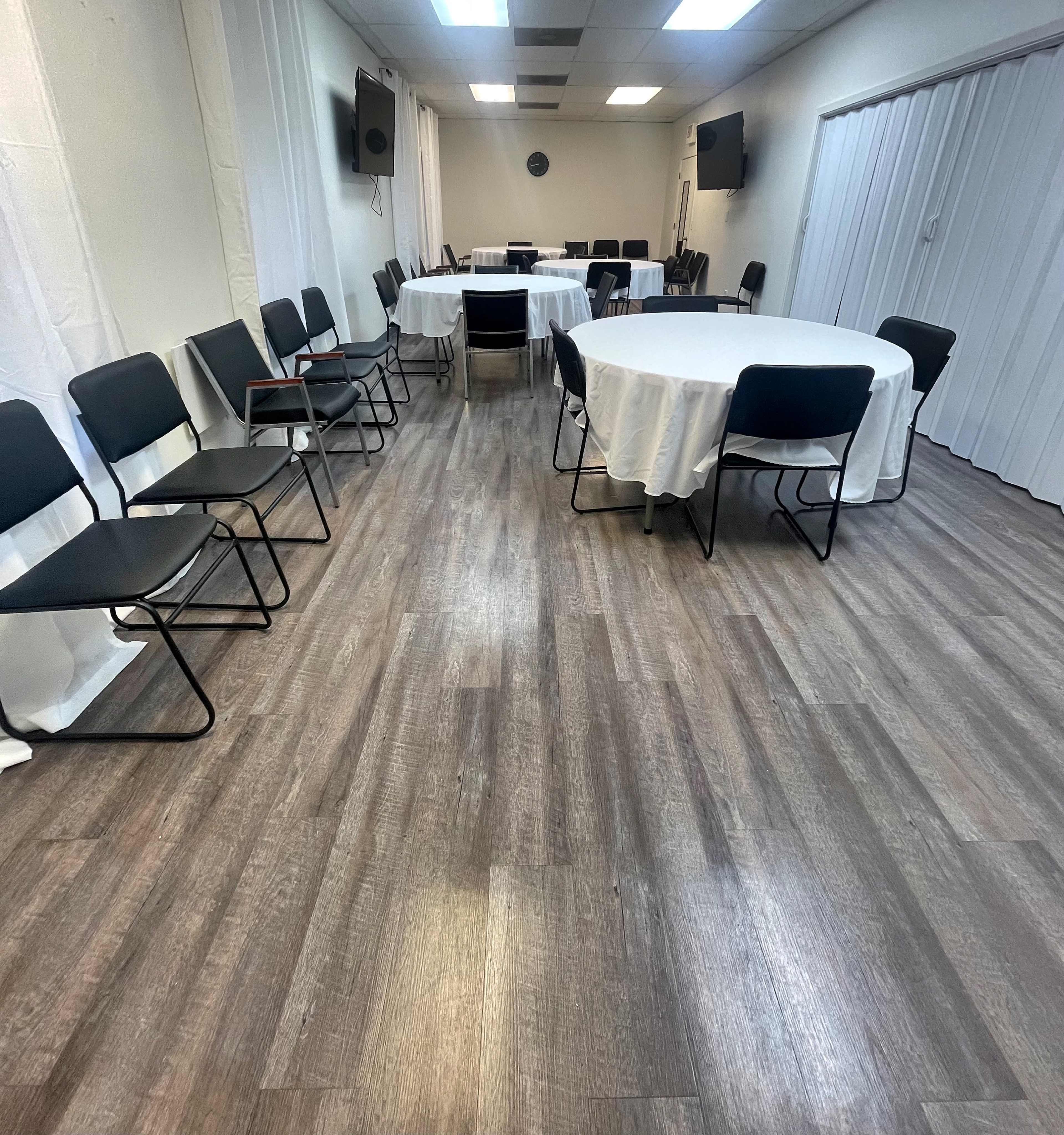 The image shows an event space with several round tables covered in white tablecloths and black folding chairs arranged around them, set on a wooden floor.