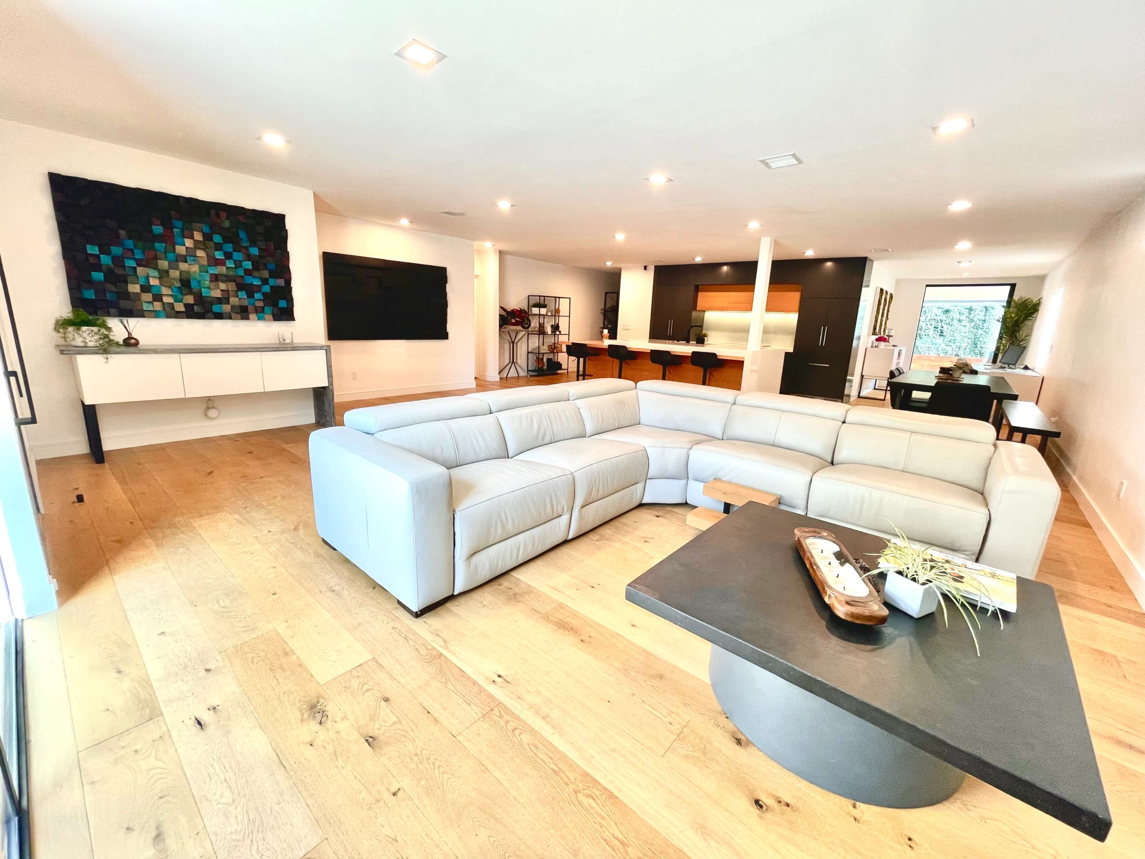 A modern living room with a light-colored sectional sofa, a black coffee table, a wall-mounted television, and an open kitchen in the background.