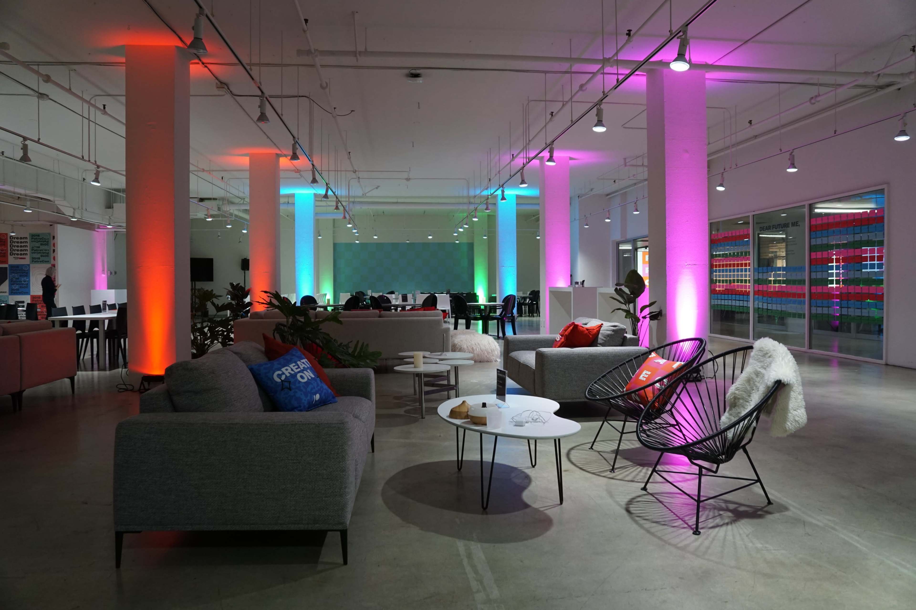 The scene features a modern, spacious lounge area with colorful lighting, comfortable seating, and a mix of tables and chairs for socializing or working.