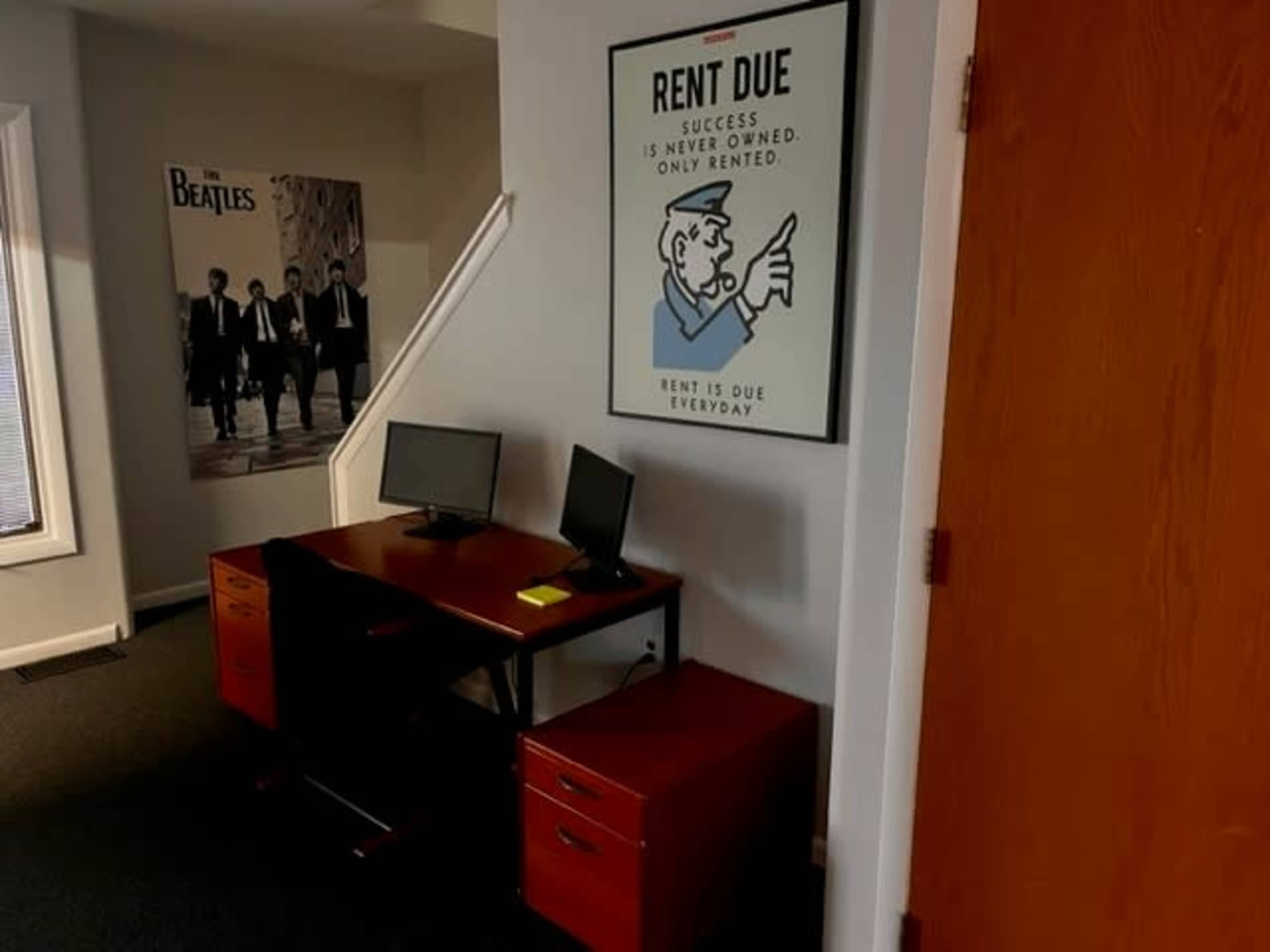 An office space with two computers on a wooden desk, a motivational poster on the wall, and a staircase in the background.