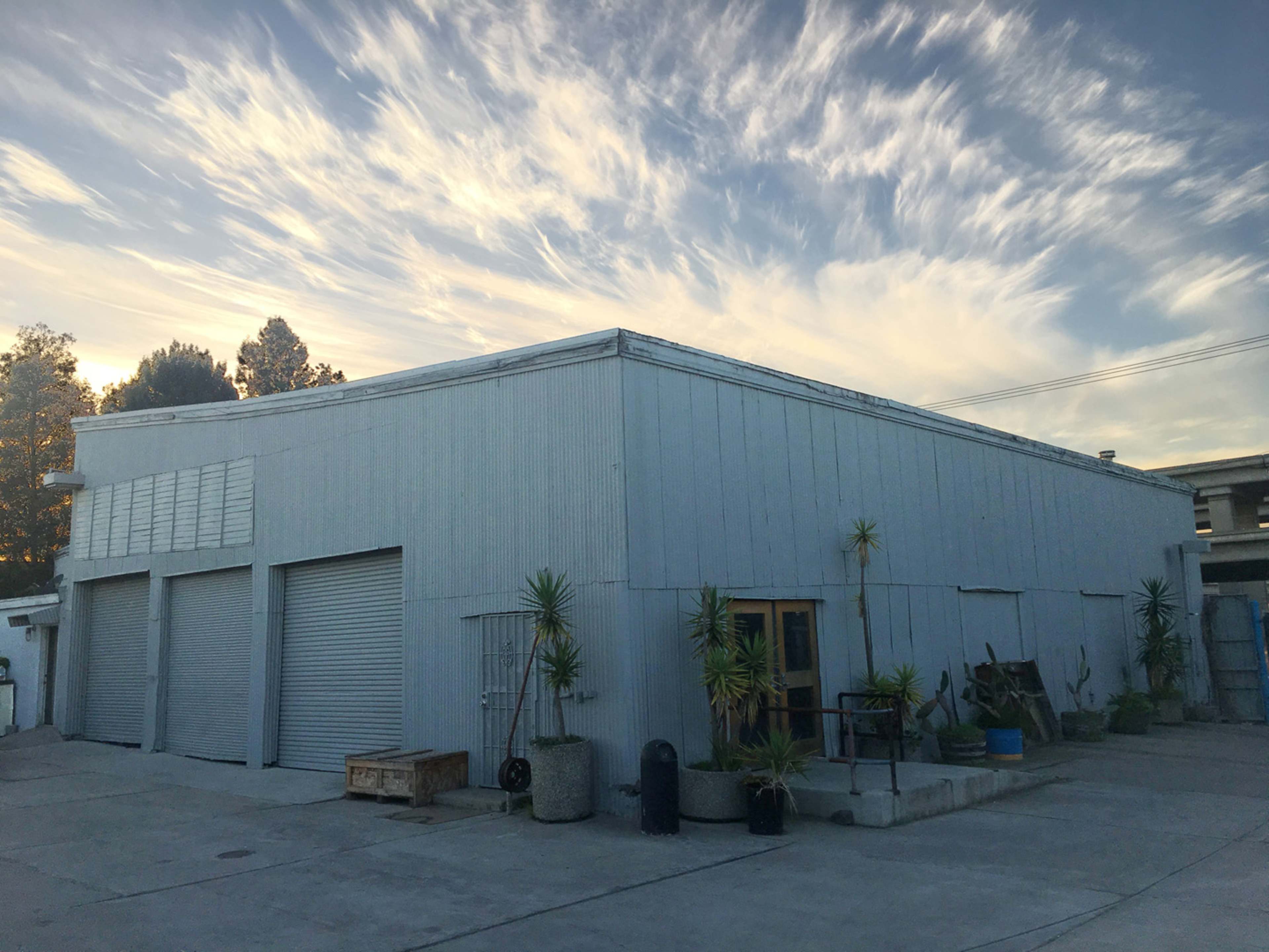 A gray, industrial building with roll-up doors sits under a sky filled with wispy clouds.
