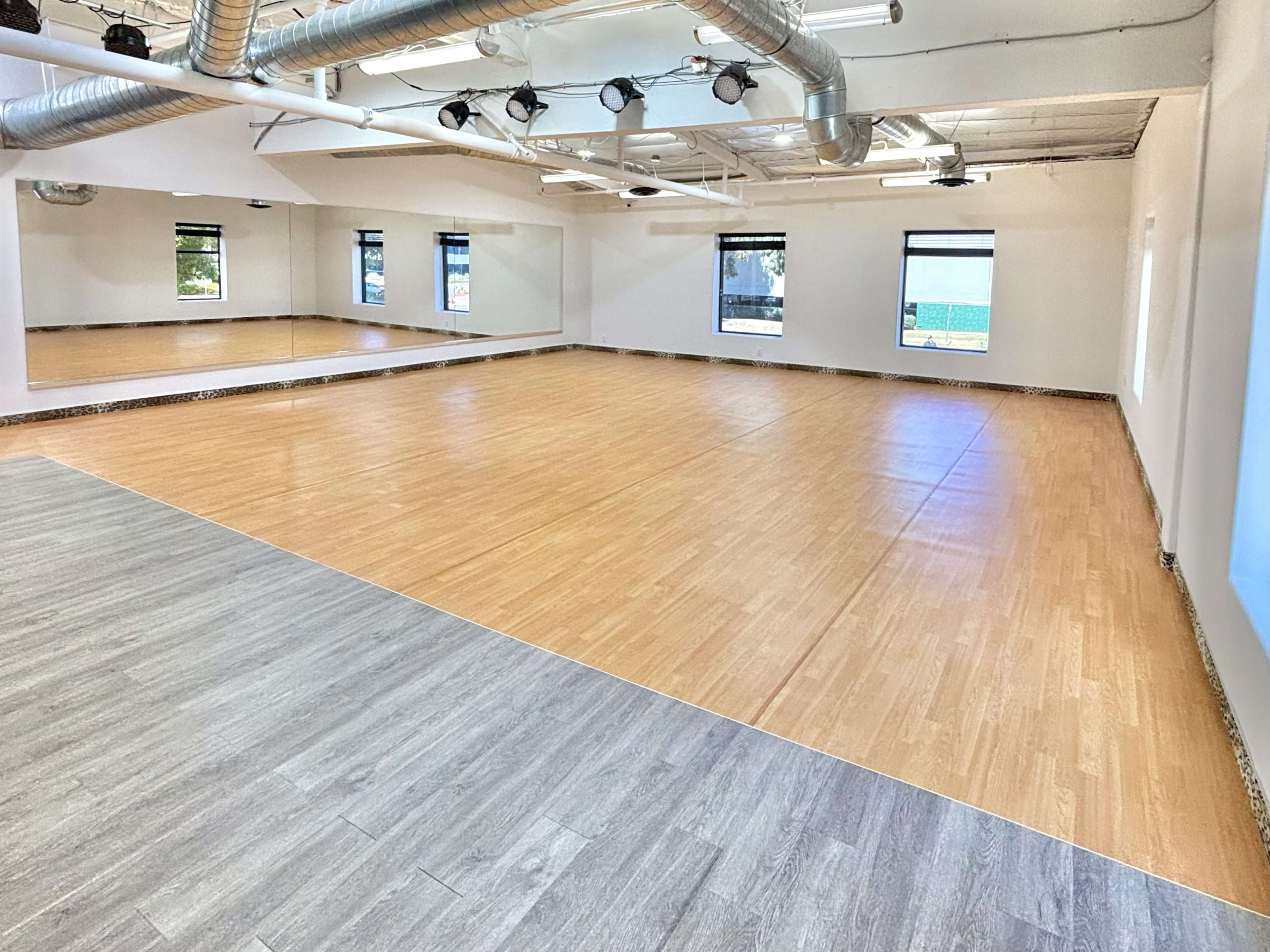 The image shows a spacious room with wooden flooring and large windows, featuring mirrored walls and overhead lighting.