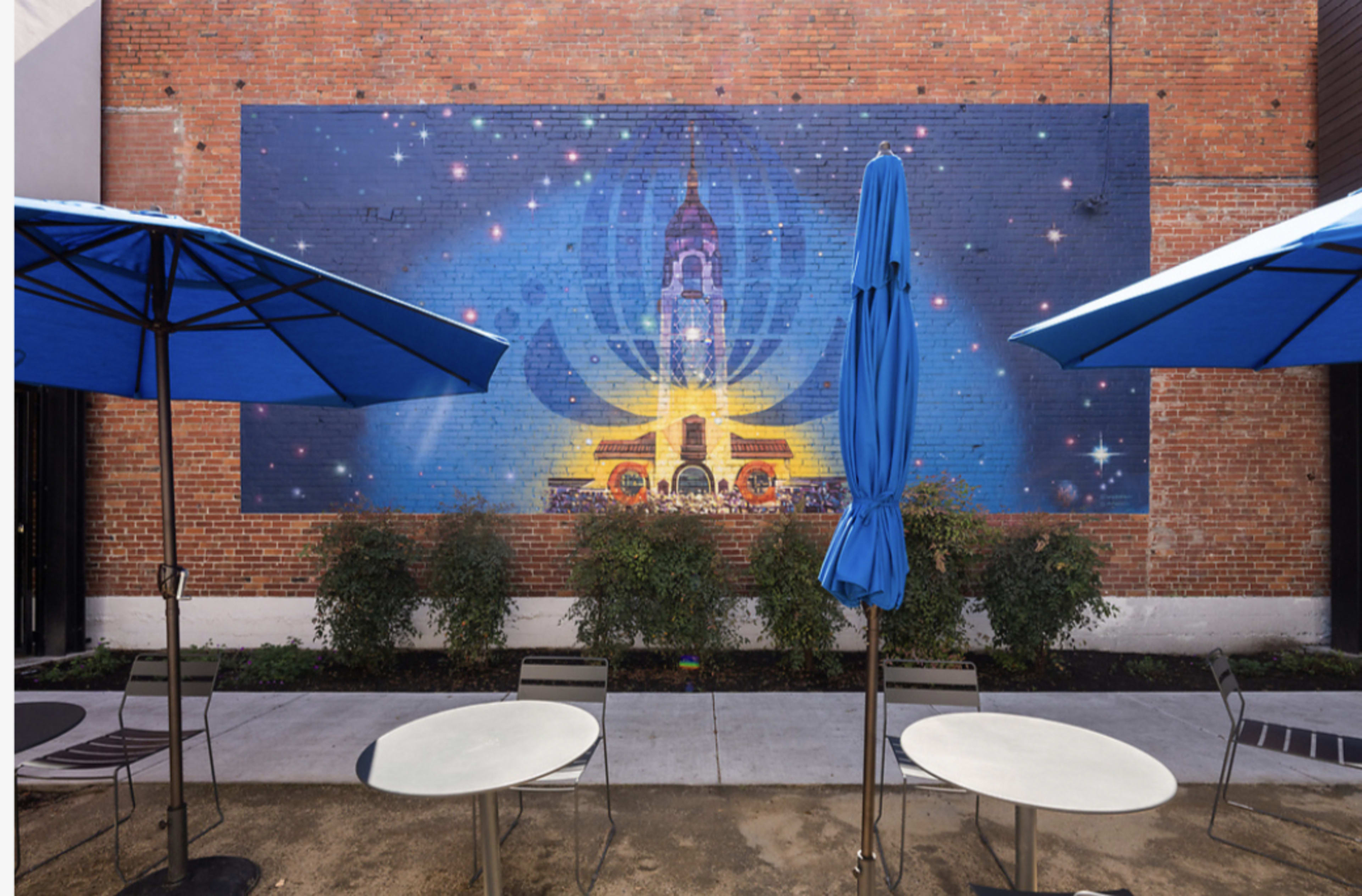 A colorful mural depicting a tower with surrounding stars is painted on a brick wall, framed by blue umbrellas and outdoor tables.