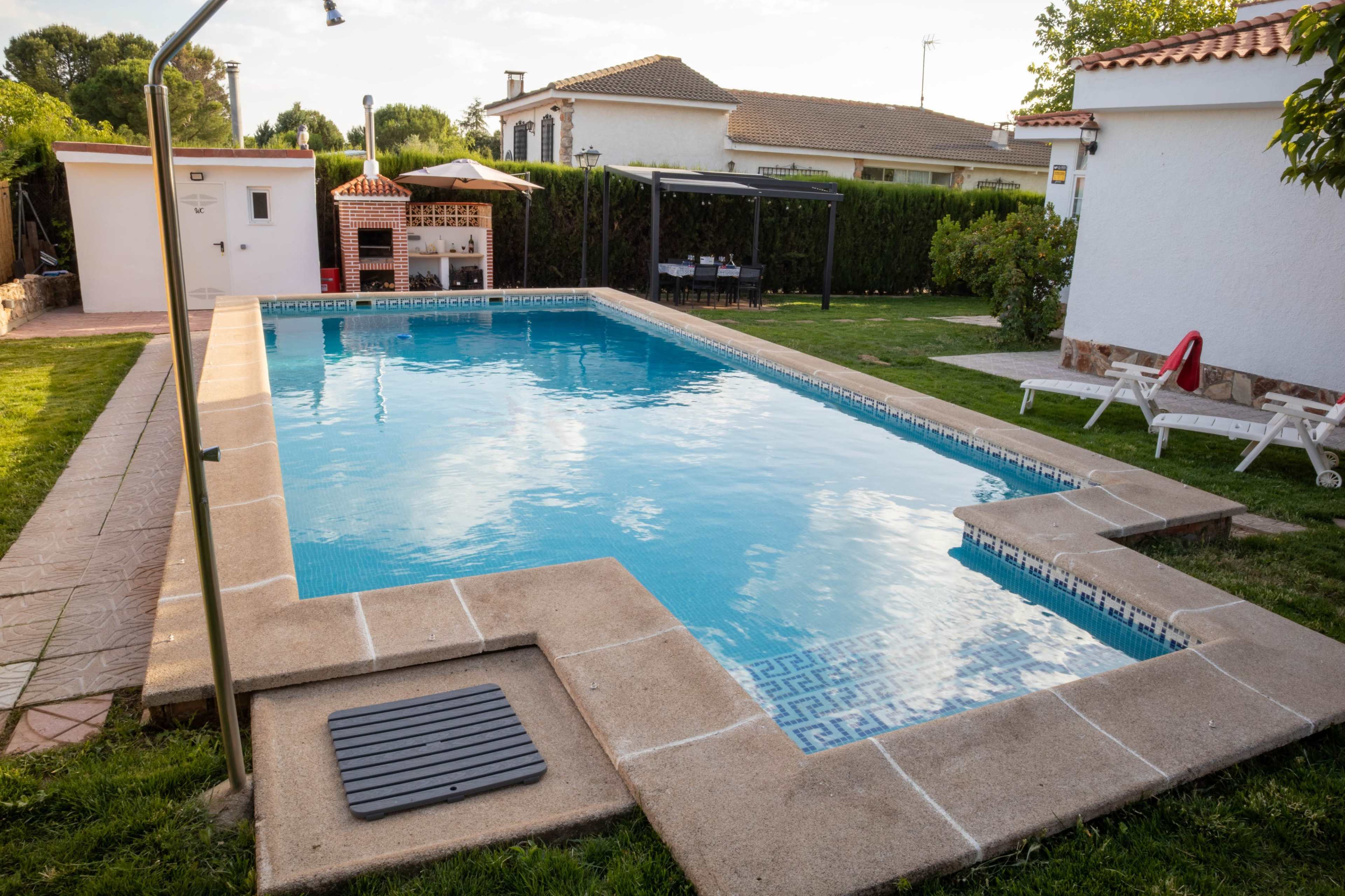 The image shows a clean swimming pool surrounded by grassy areas and a few structures, including a small outdoor kitchen and seating area.