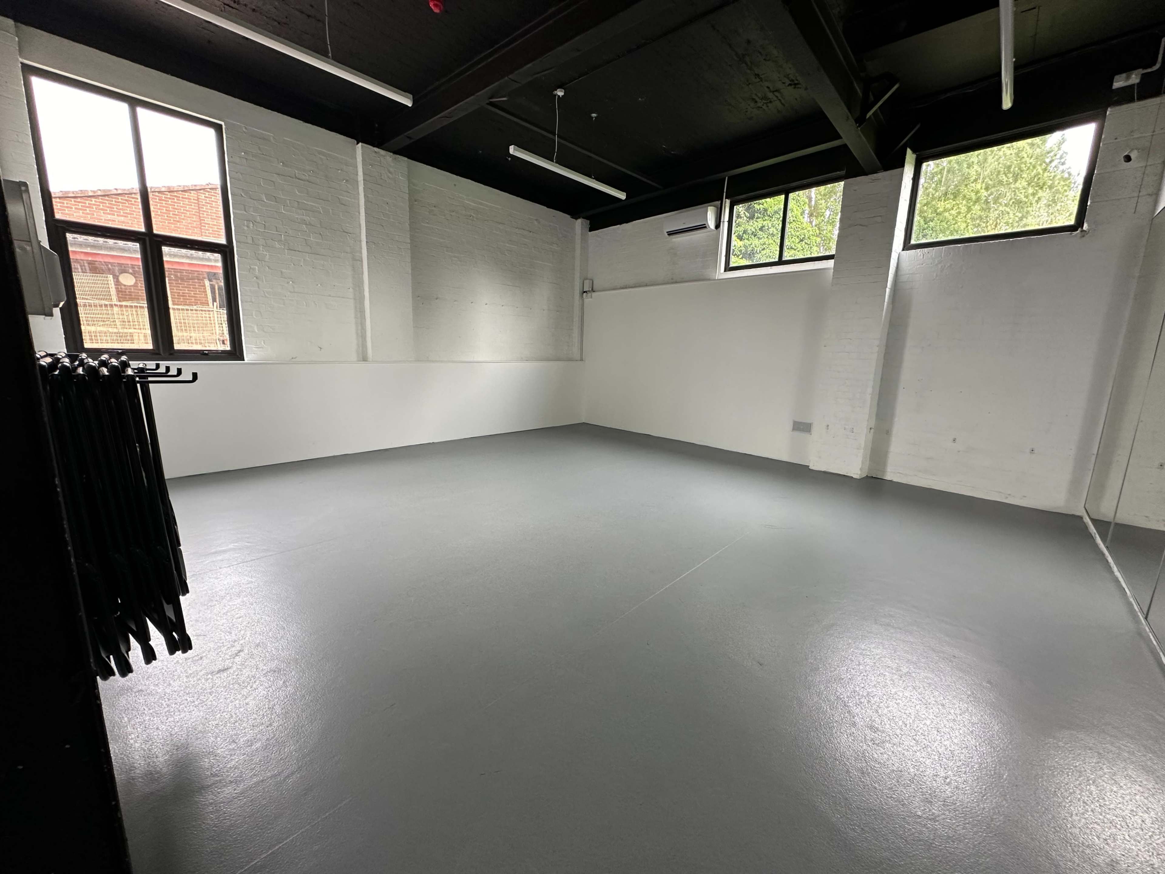 An empty room with gray flooring and large windows, featuring a white brick wall and a clothing rack with black garments in one corner.