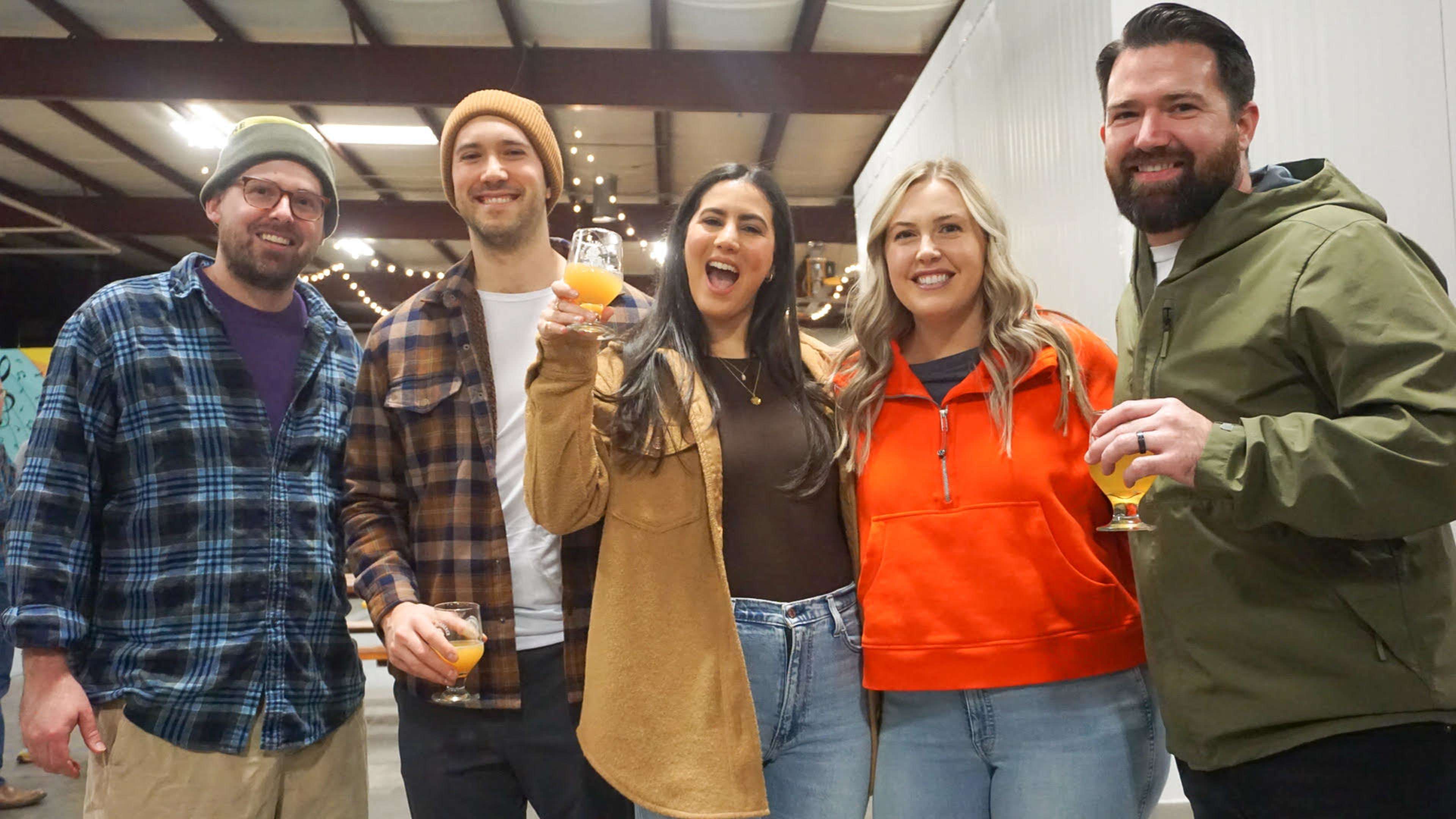 A group of five people stands together indoors, smiling and holding drinks at a social gathering.