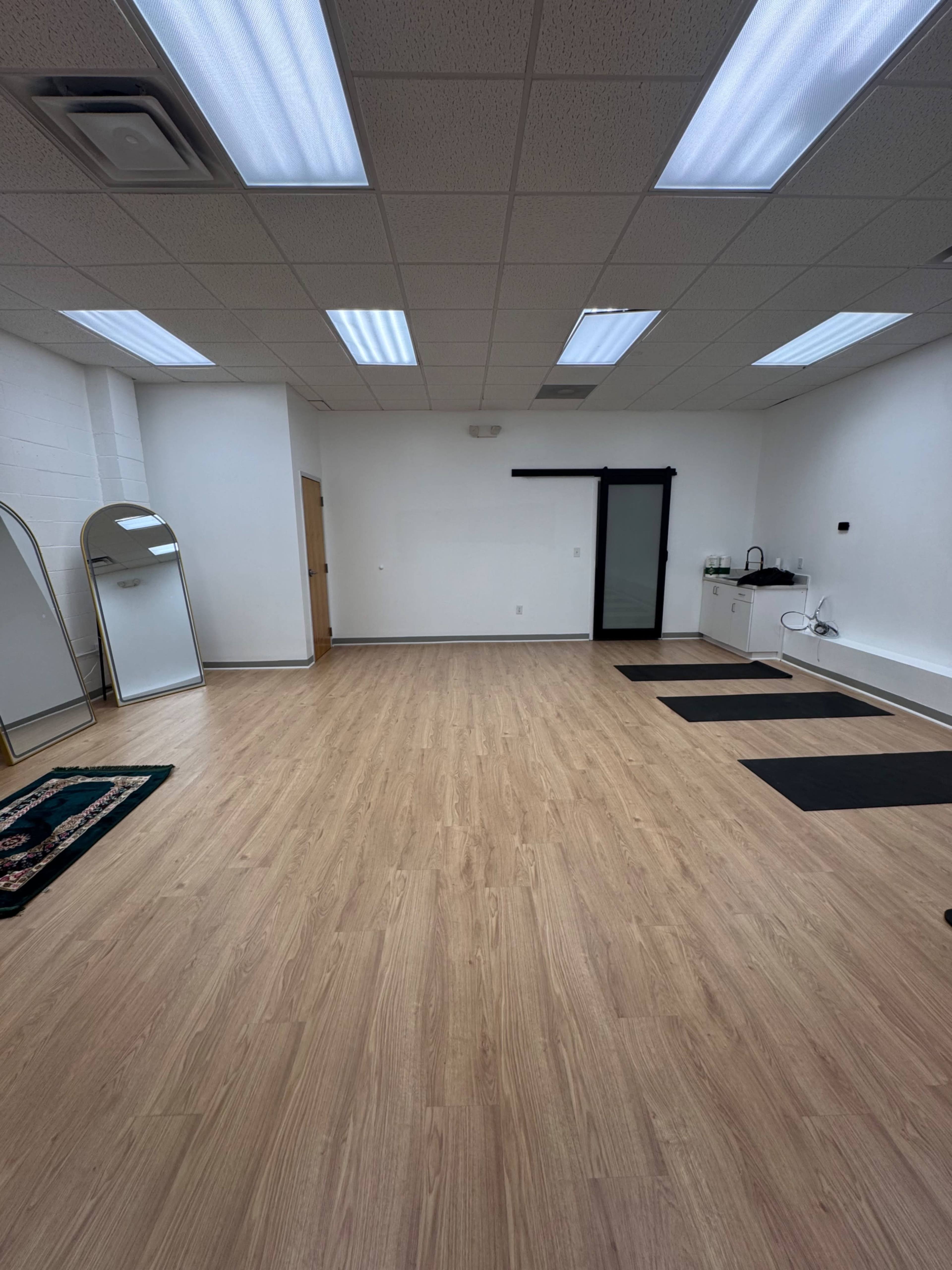 The image shows a spacious, empty room with wooden flooring, two large mirrors, and a small kitchenette area along one wall.