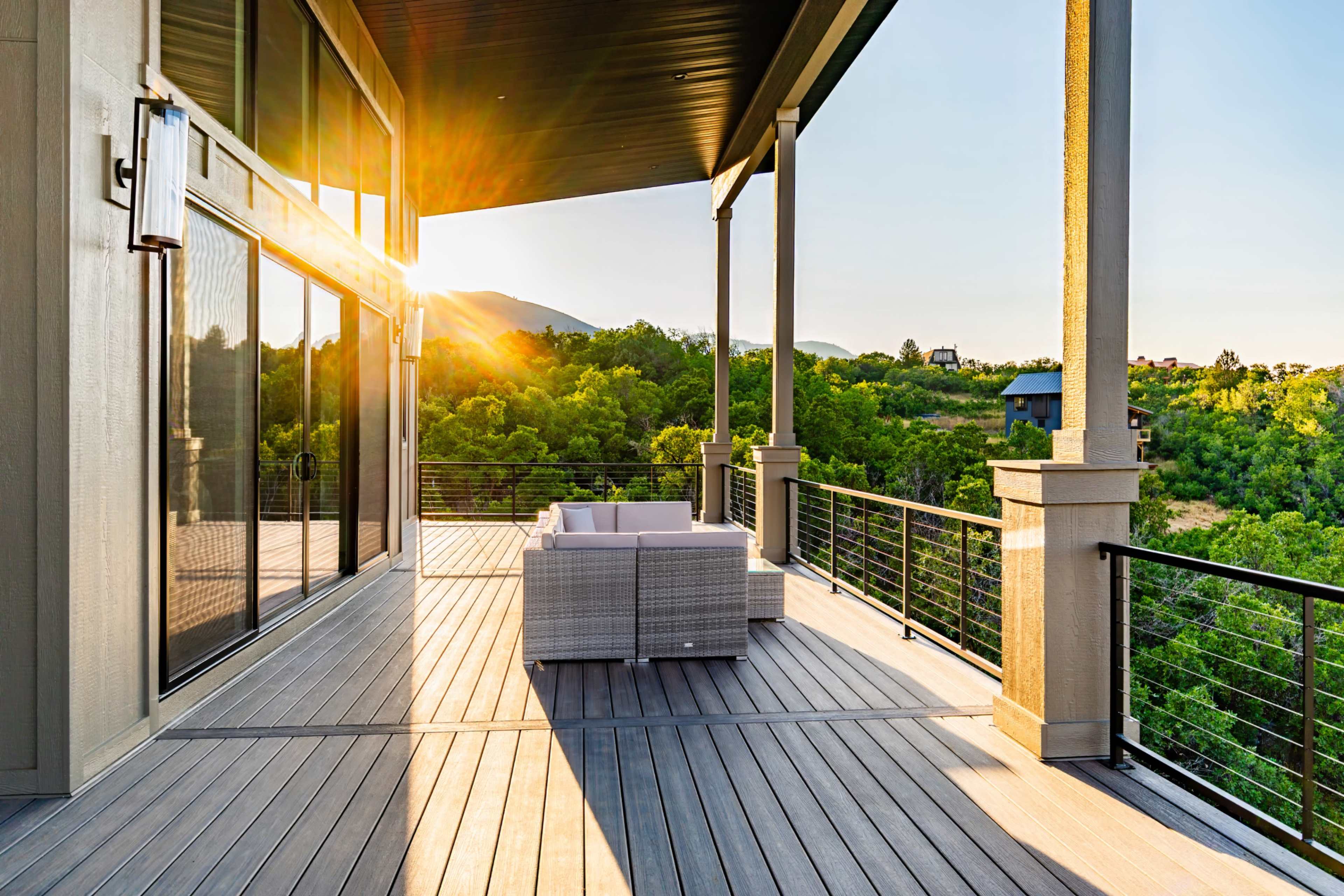 The image shows a spacious outdoor deck with a modern seating area, framed by a scenic view of green hills and a setting sun.