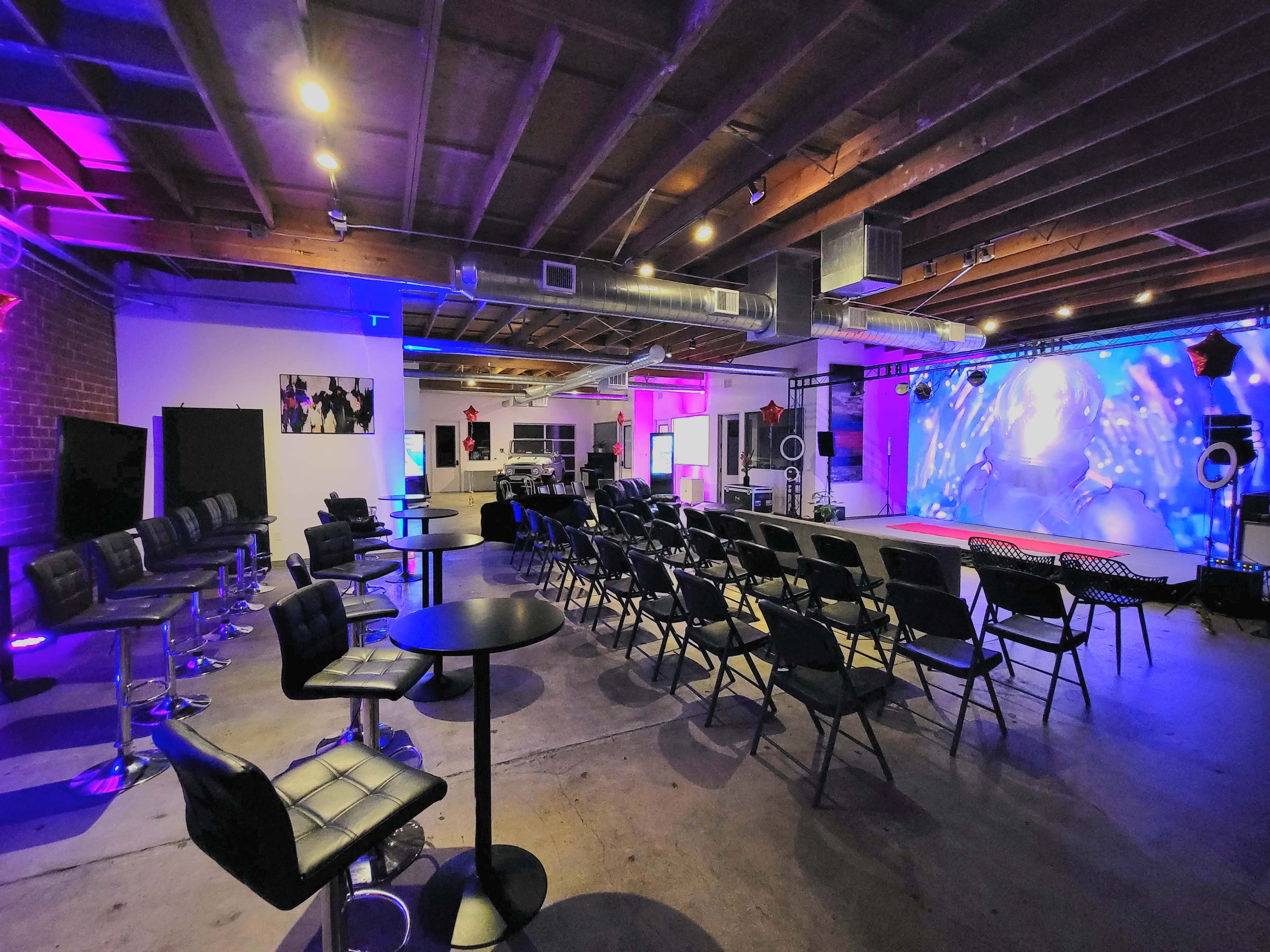 The image shows a spacious event venue with black chairs arranged in rows, small tables, and colorful lighting along the walls and stage area.