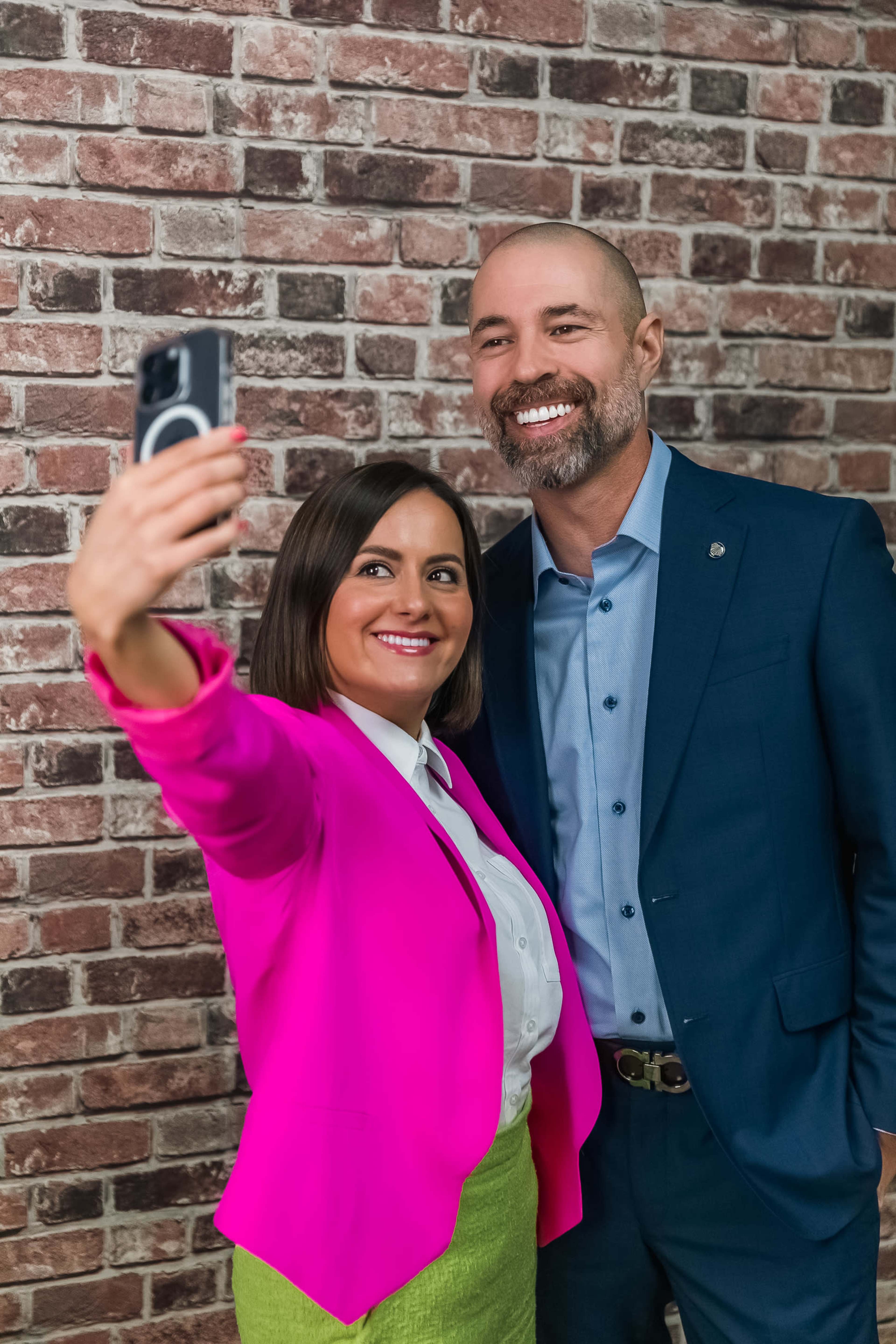 A woman in a pink blazer and a man in a blue suit are posing together for a selfie in front of a brick wall.