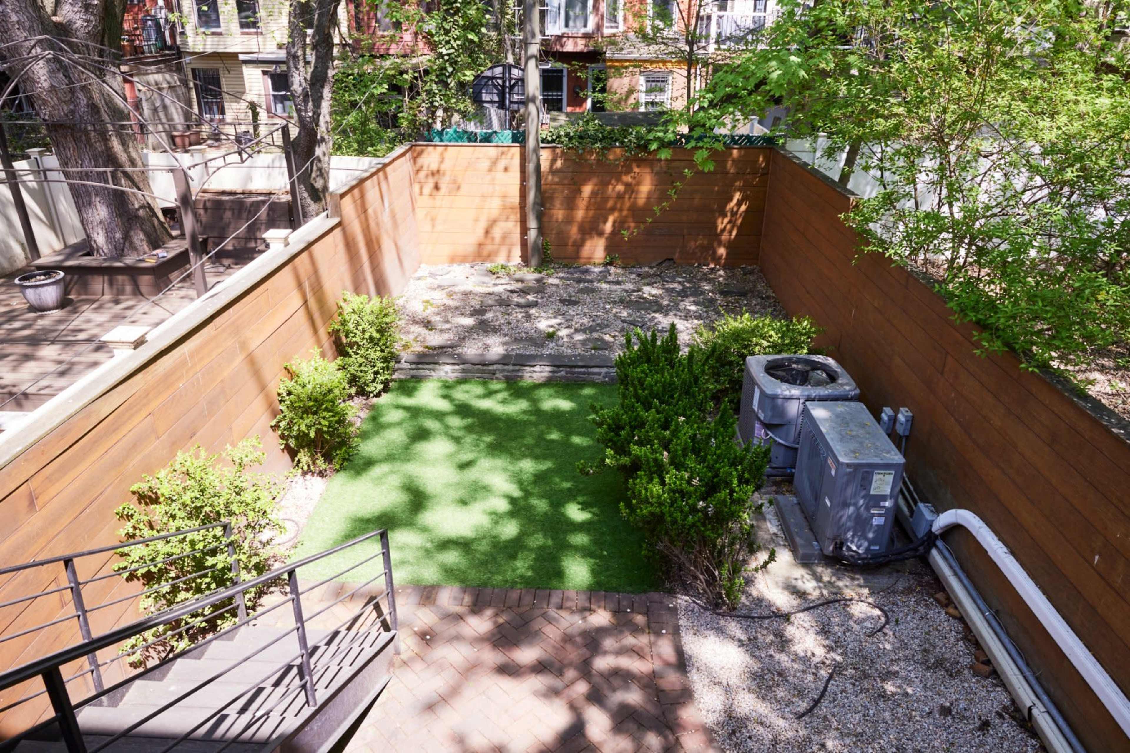 The image shows a small backyard enclosed by wooden fences, featuring a patch of artificial turf, a gravel area, and an air conditioning unit.