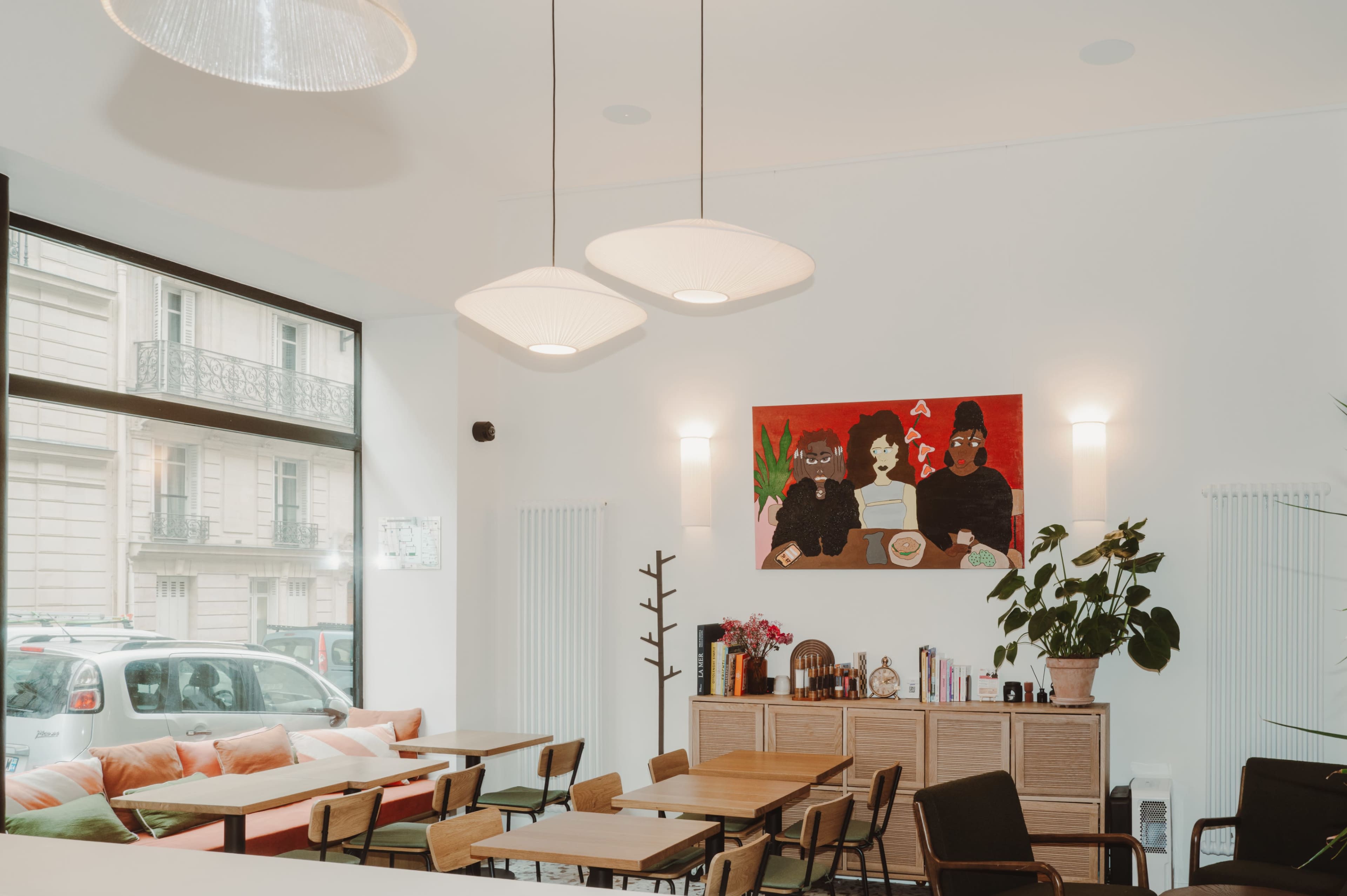 The image depicts a modern café interior with wooden tables, colorful seating, a large painting on the wall, and soft pendant lighting.