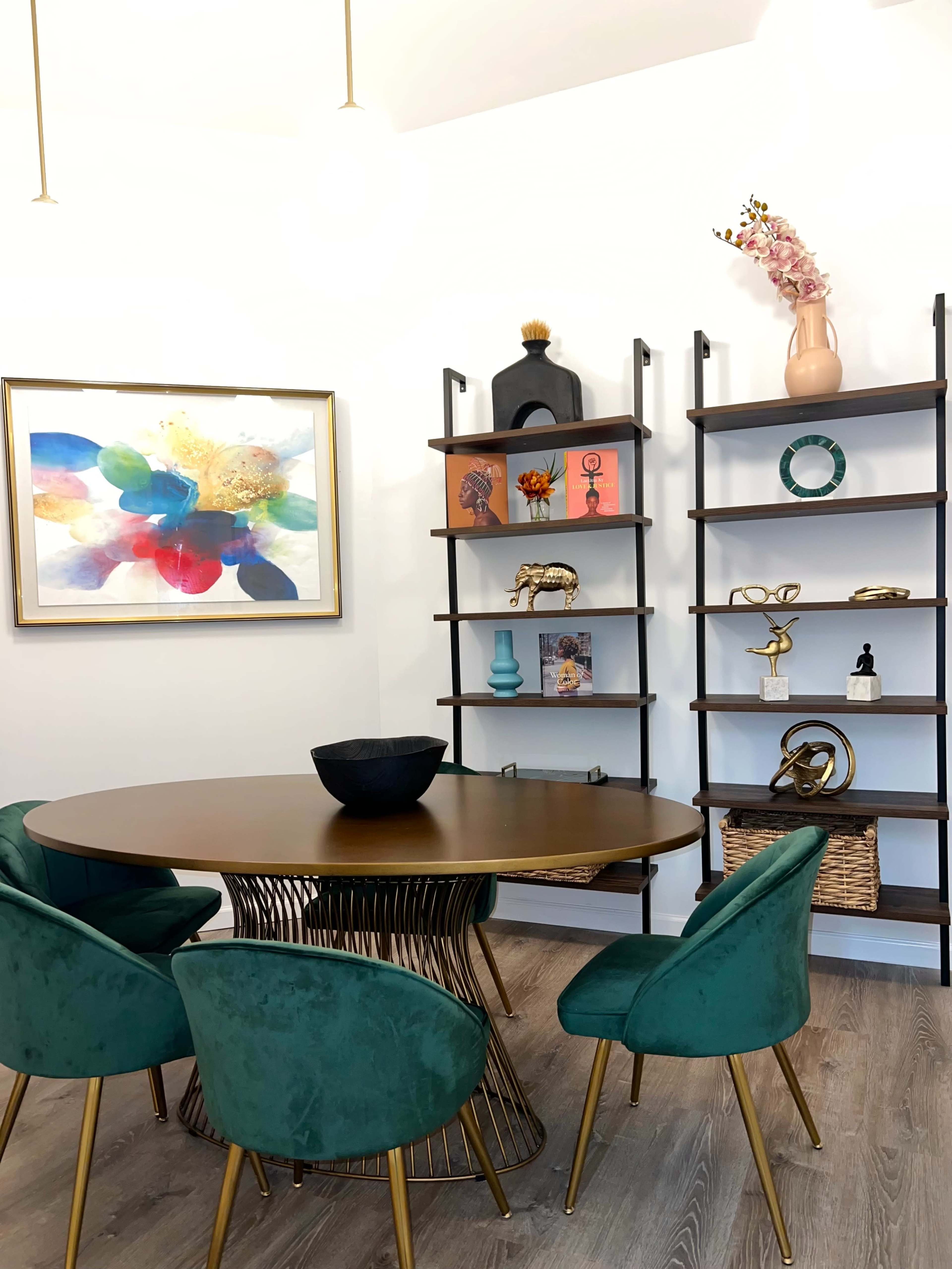 A modern dining area with a round table surrounded by green upholstered chairs, a colorful artwork on the wall, and two open shelves displaying decorative items.