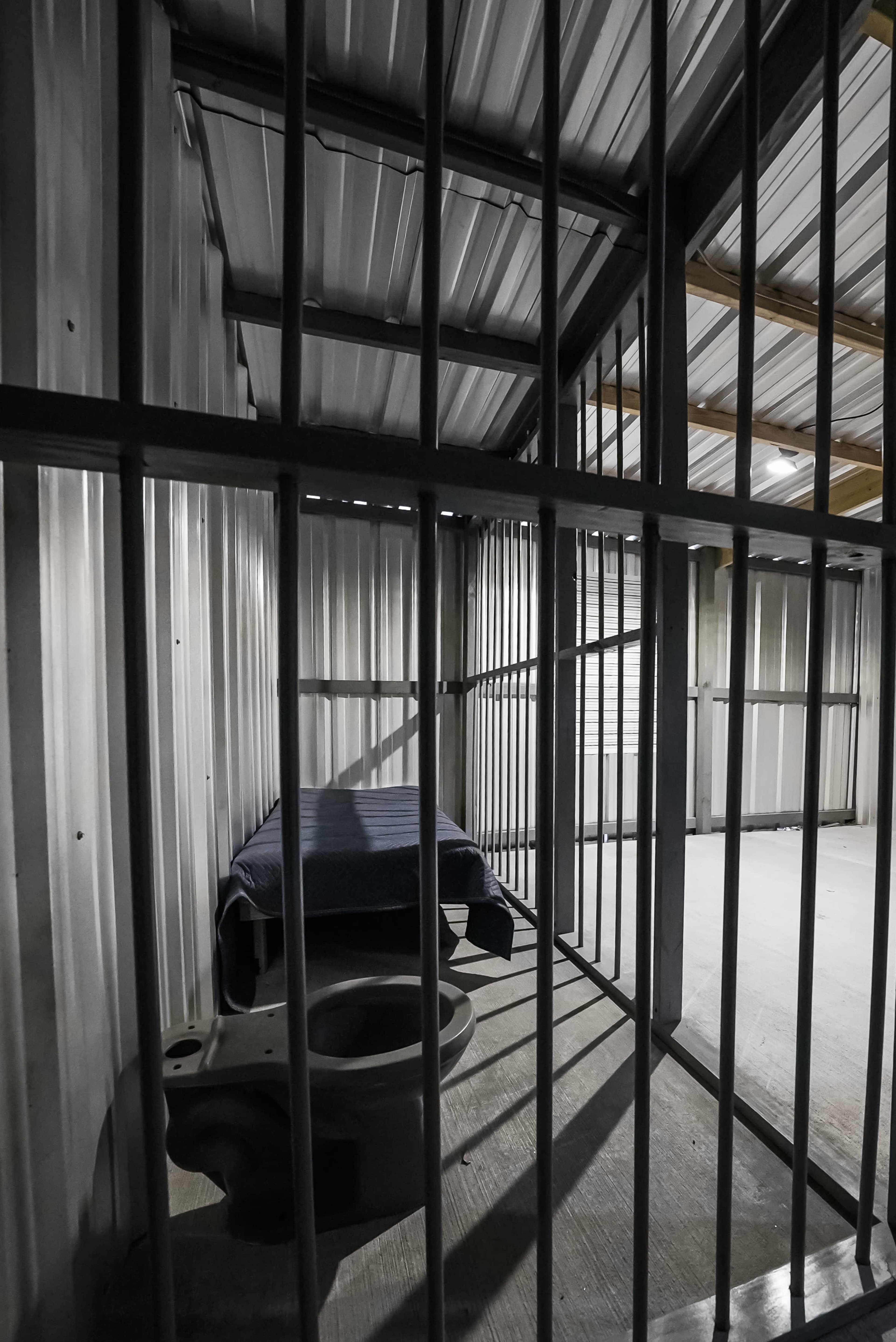 The image shows a dimly lit confinement cell with metal bars, a bed covered with a blue blanket, and a toilet fixture.