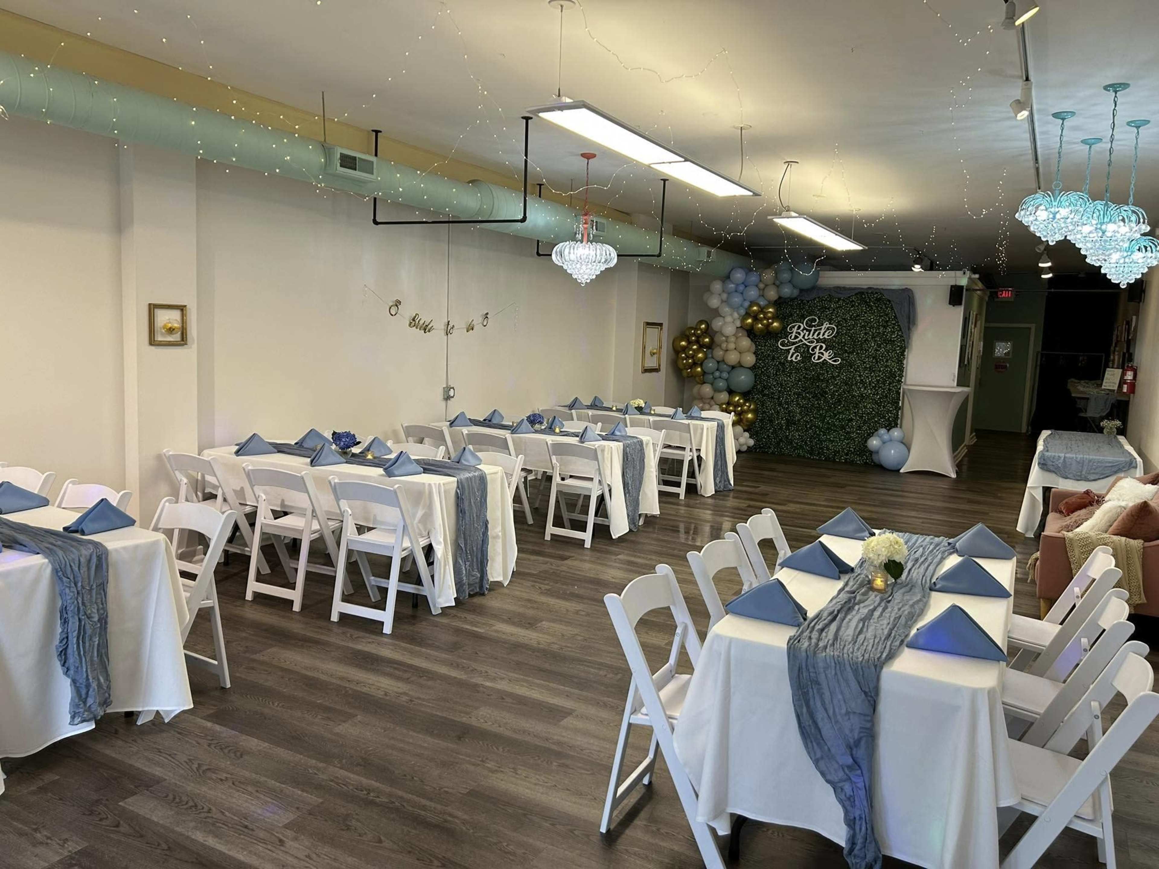 A decorated event space features neatly arranged tables with white linens and blue napkins, illuminated by hanging lights and adorned with balloons.
