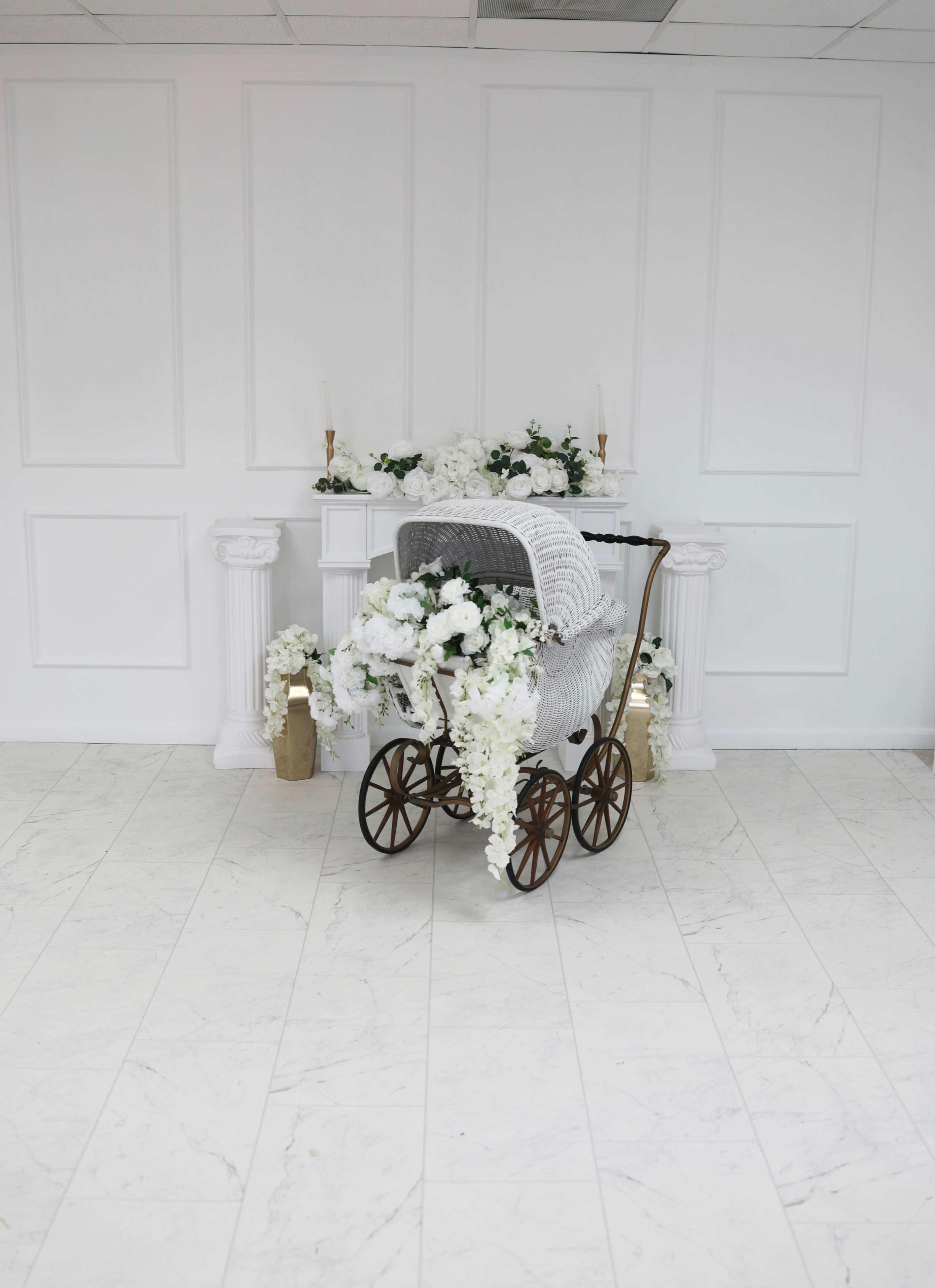 A vintage baby carriage adorned with white flowers is positioned in front of a decorative white mantel in a bright, minimalist room.