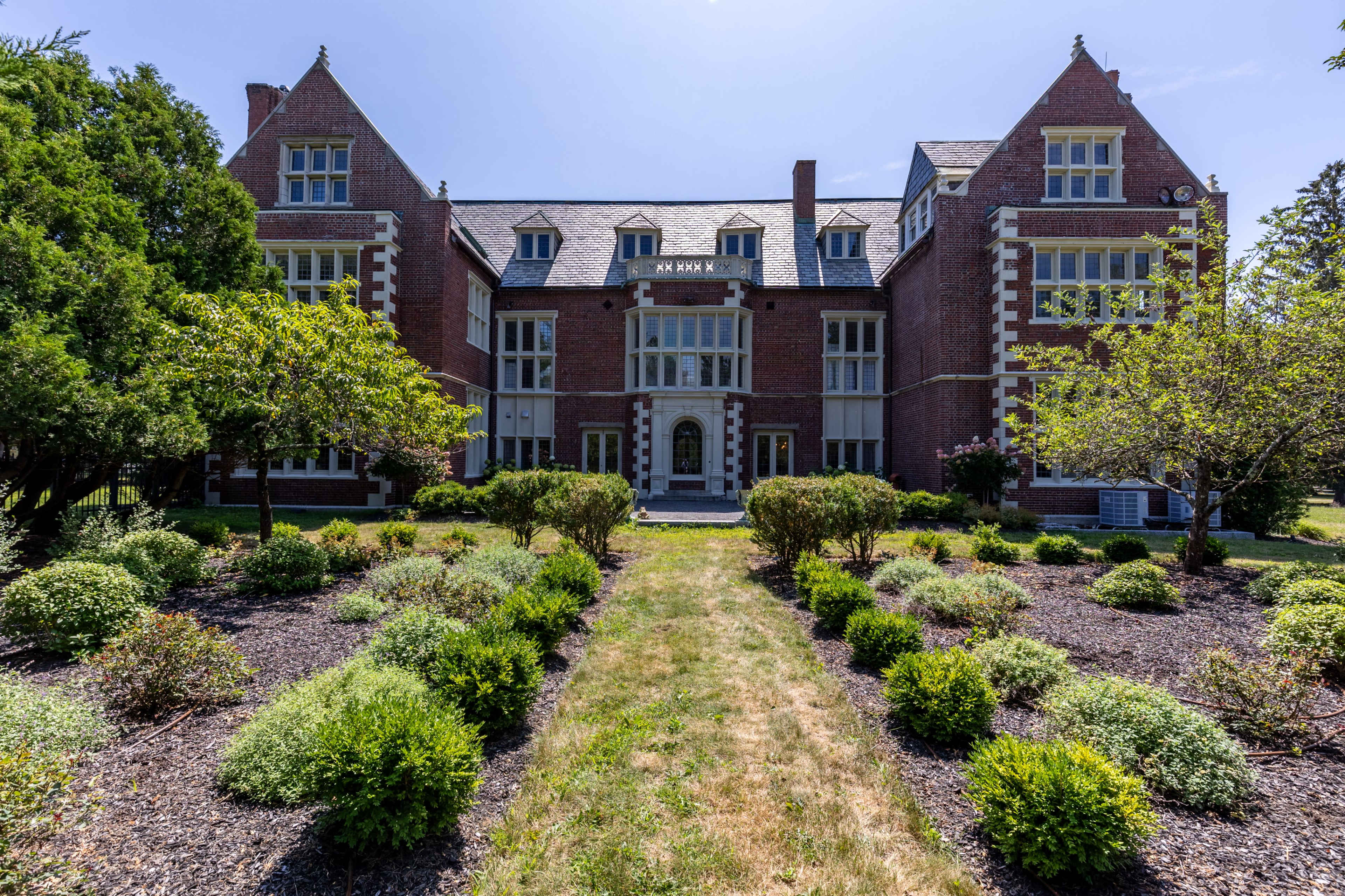 The image shows a large brick mansion with multiple gabled roofs, flanked by well-kept gardens and trees in the foreground.
