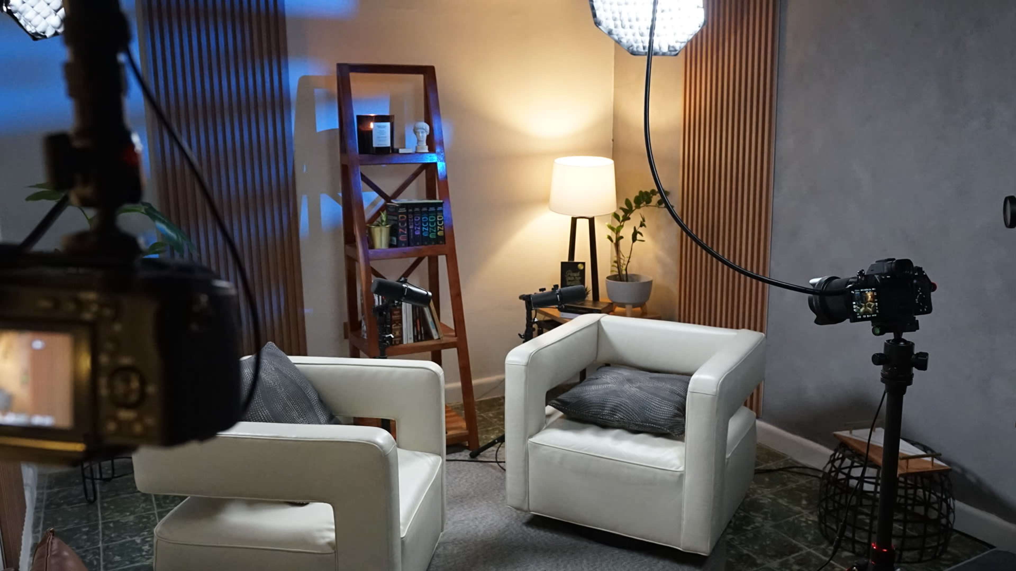 The image shows a cozy indoor setup with two white armchairs, a small table, a plant, a lamp, and various cameras positioned for filming.