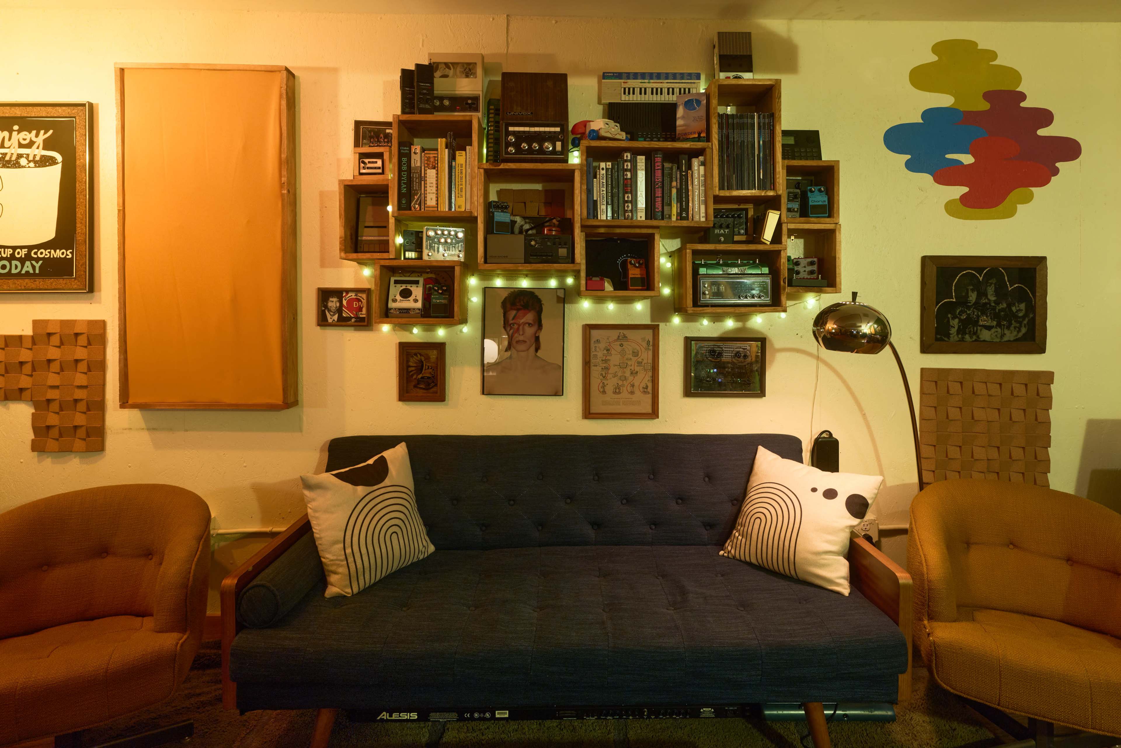 The image shows a cozy living space featuring a blue sofa with patterned cushions, framed artwork on the walls, and shelves filled with books and decorative items.