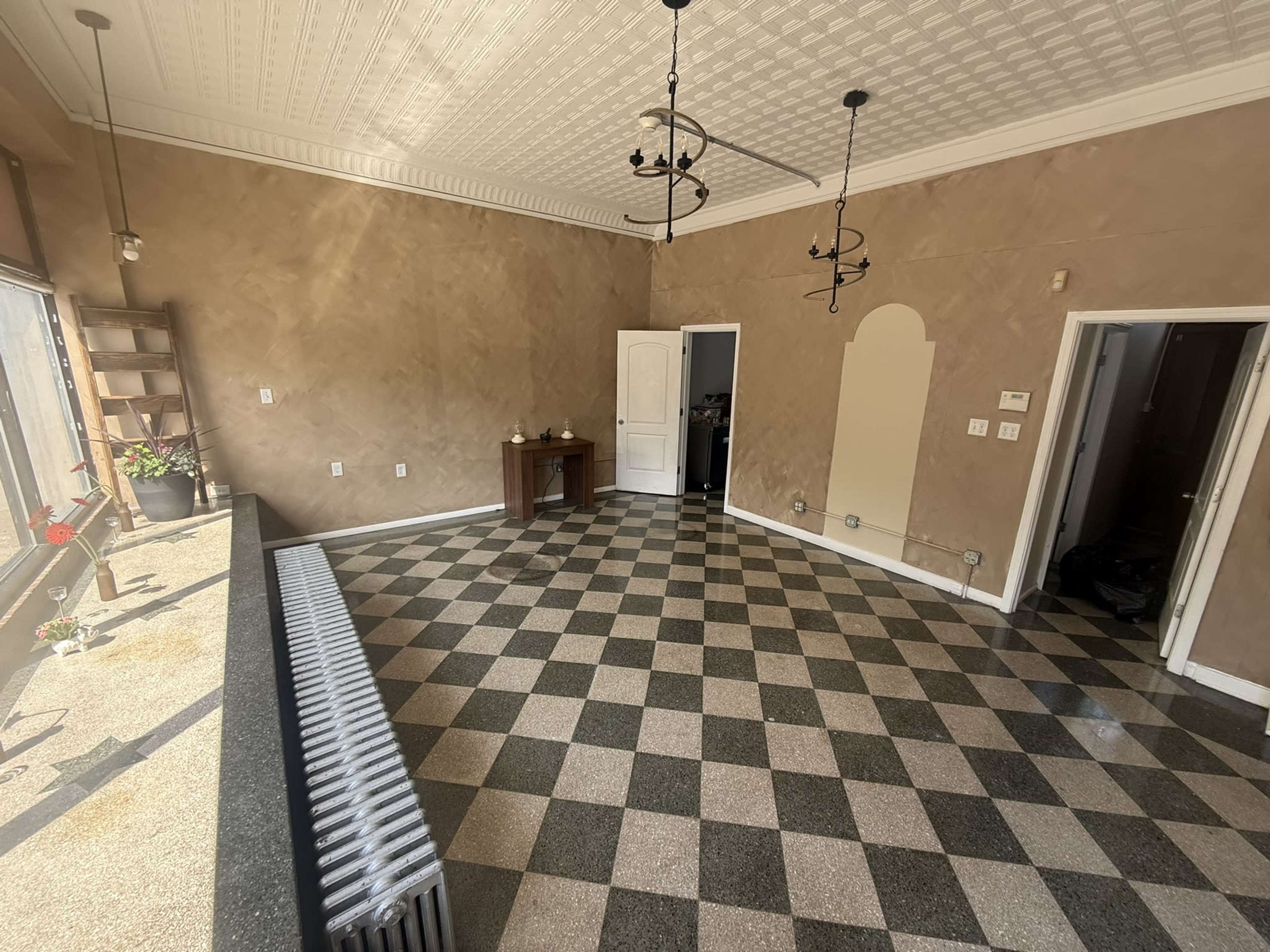 The image shows an empty room with checkered tile flooring, beige walls, and a door leading to another space.