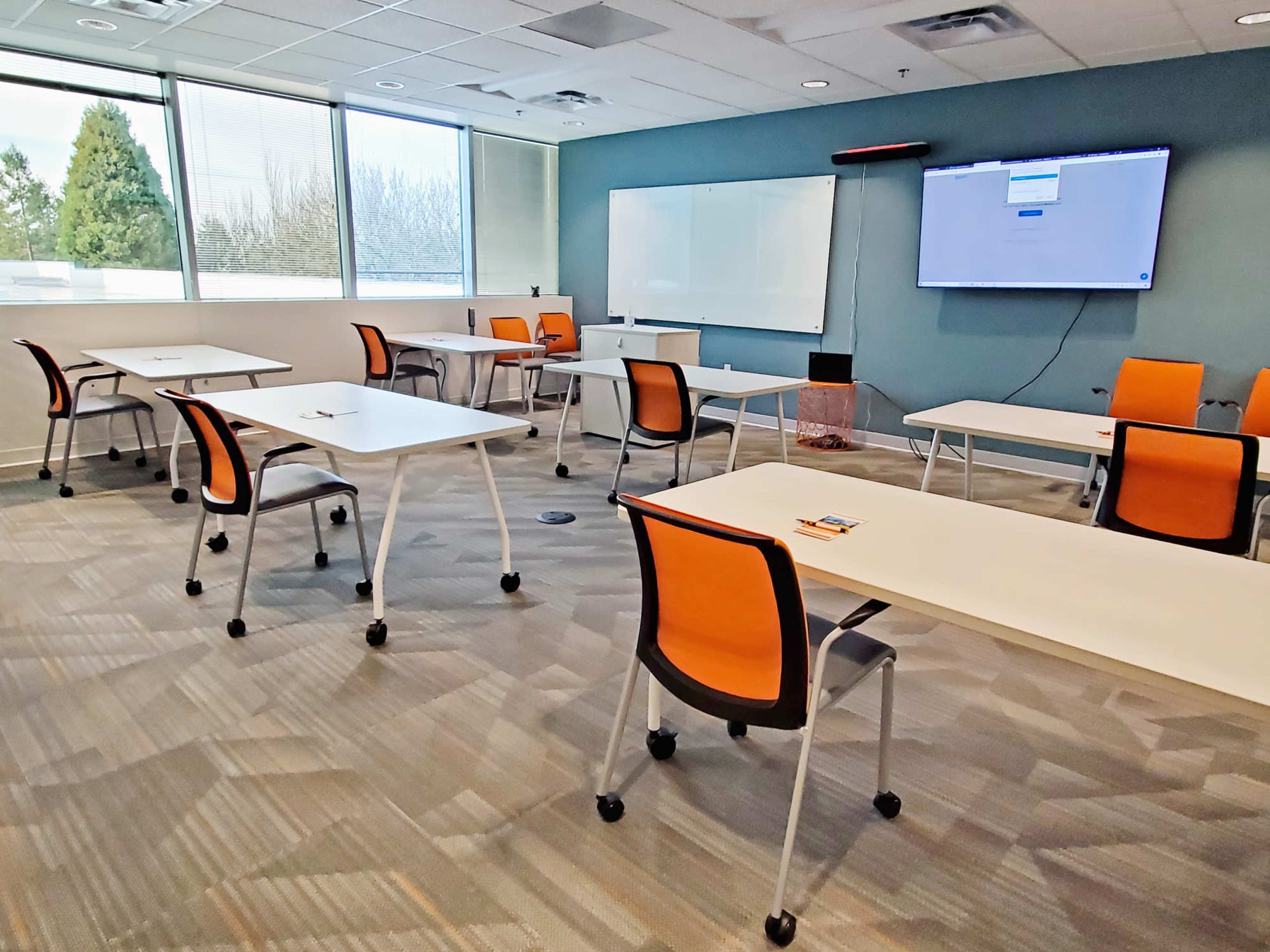 A classroom is arranged with several empty tables and orange chairs, featuring a large screen on one wall and large windows overlooking trees.