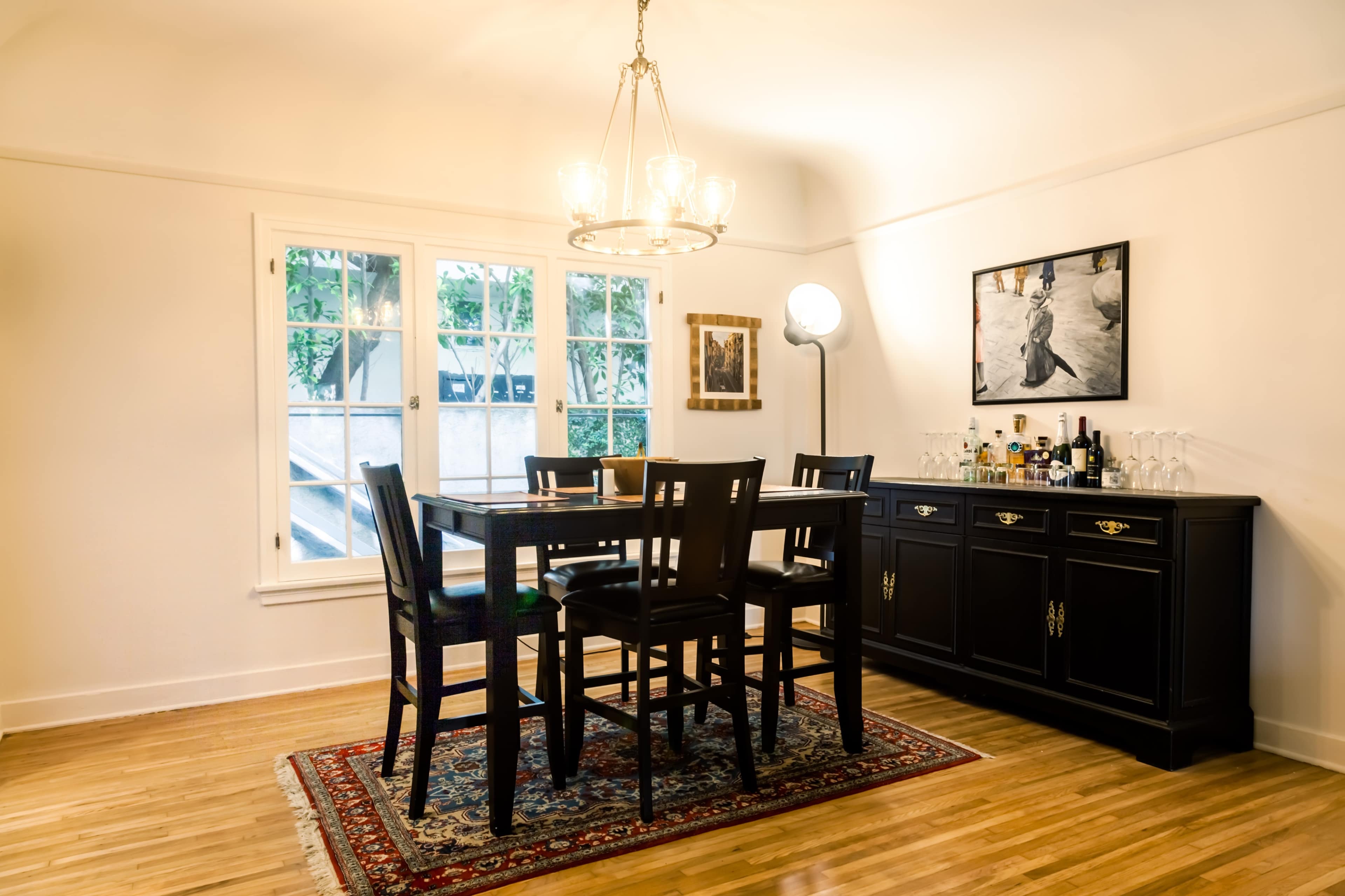 A dining area features a wooden table with high-backed chairs, a buffet cabinet adorned with bottles and glasses, and a large window providing natural light.