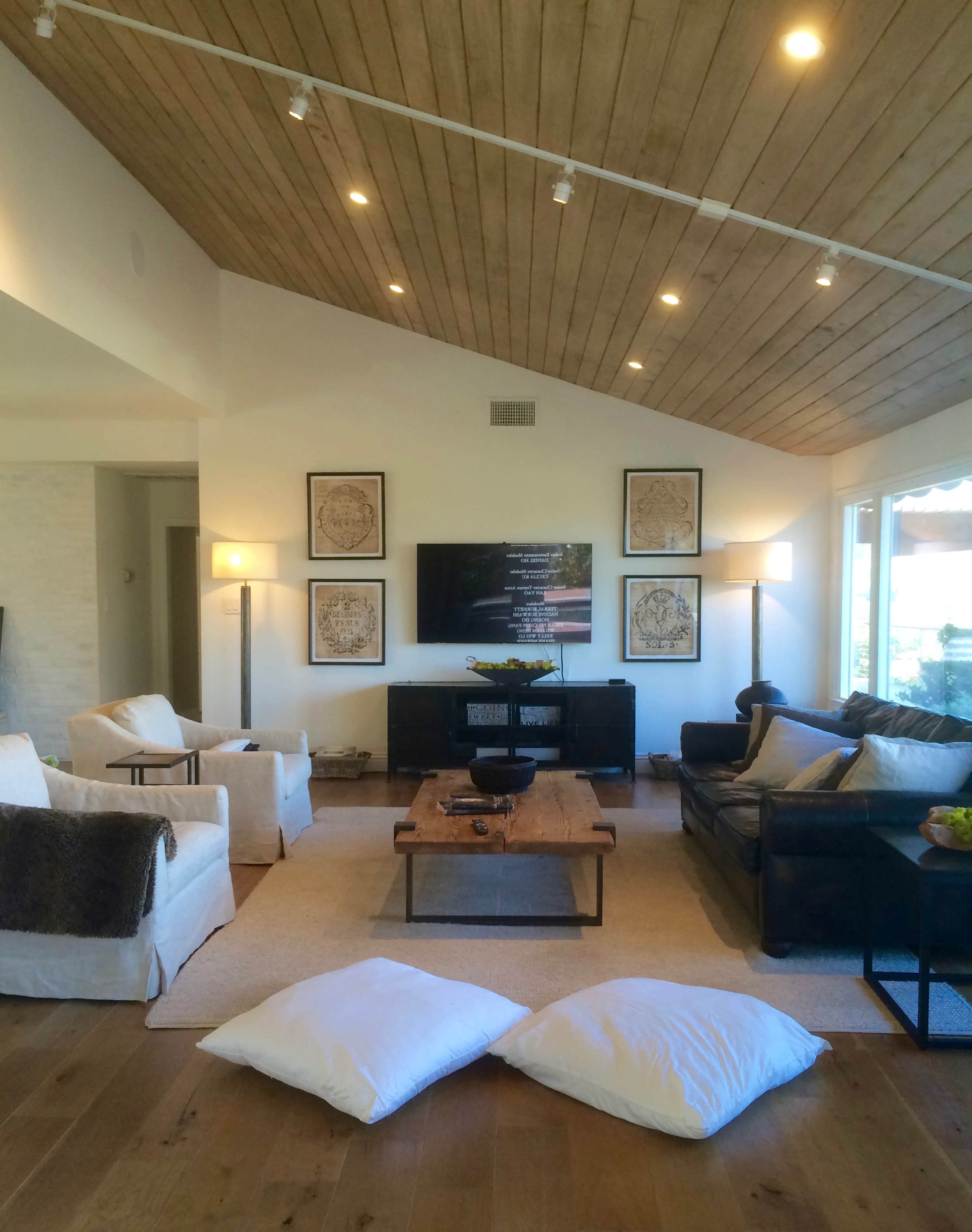 A modern living room with a high wooden ceiling, a flat-screen TV mounted on the wall, two armchairs, a leather sofa, and a wooden coffee table surrounded by decorative pillows.
