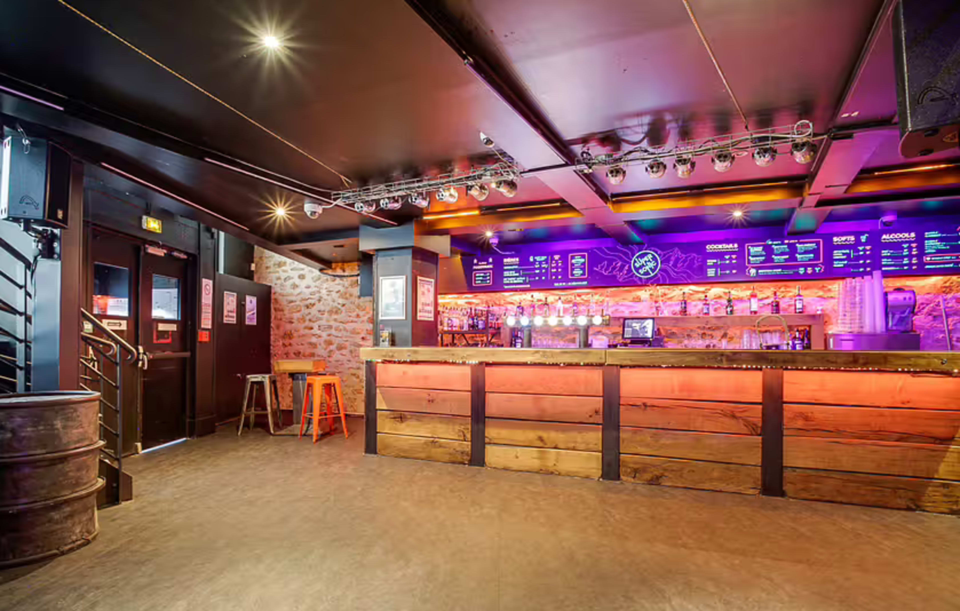 The image shows a dimly lit bar with a wooden counter, illuminated by colorful lights, and a variety of drinks displayed behind the counter.