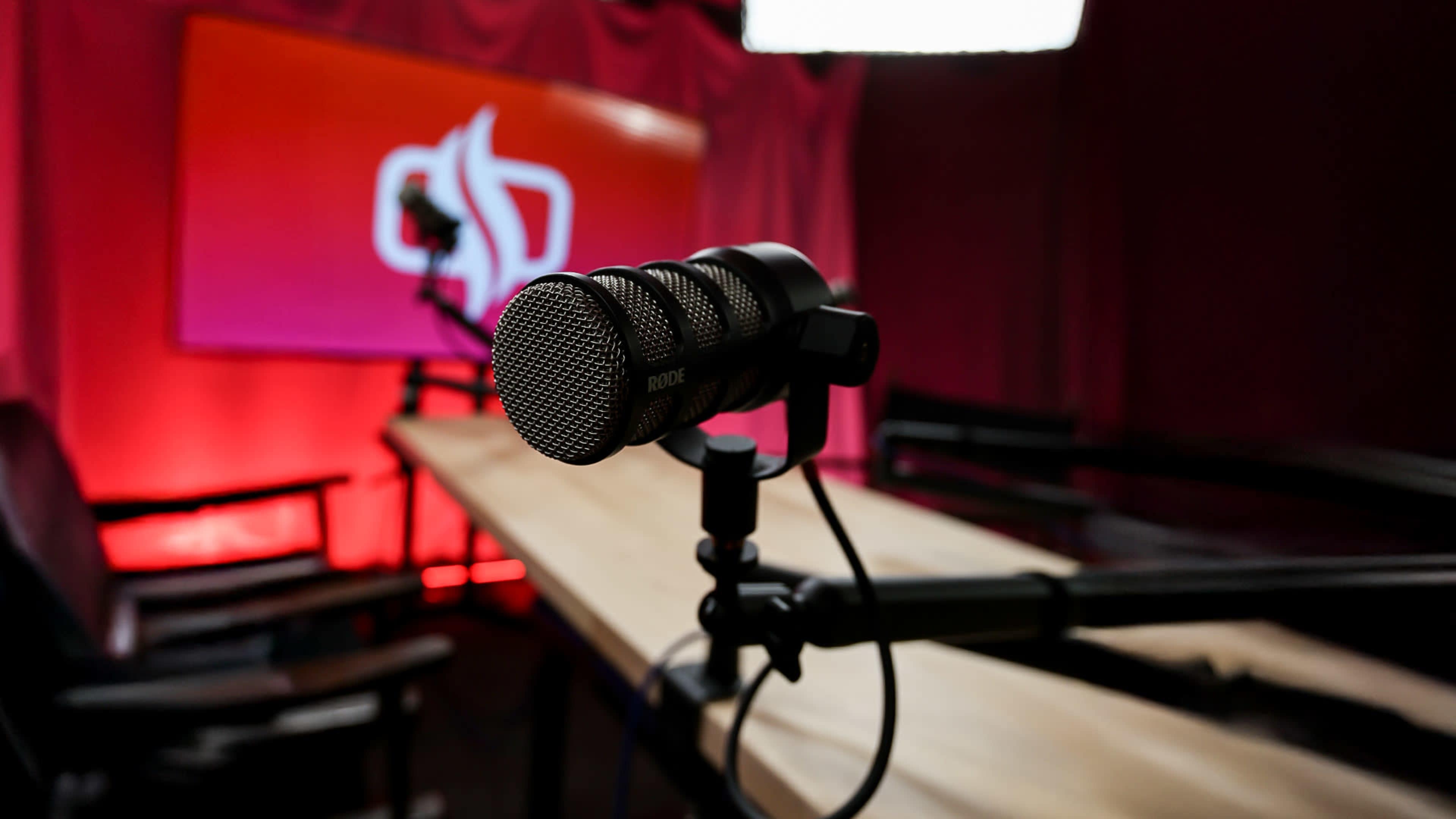 A microphone on a stand is positioned in front of a brightly lit backdrop featuring a logo, with a wooden table and chairs visible in the scene.