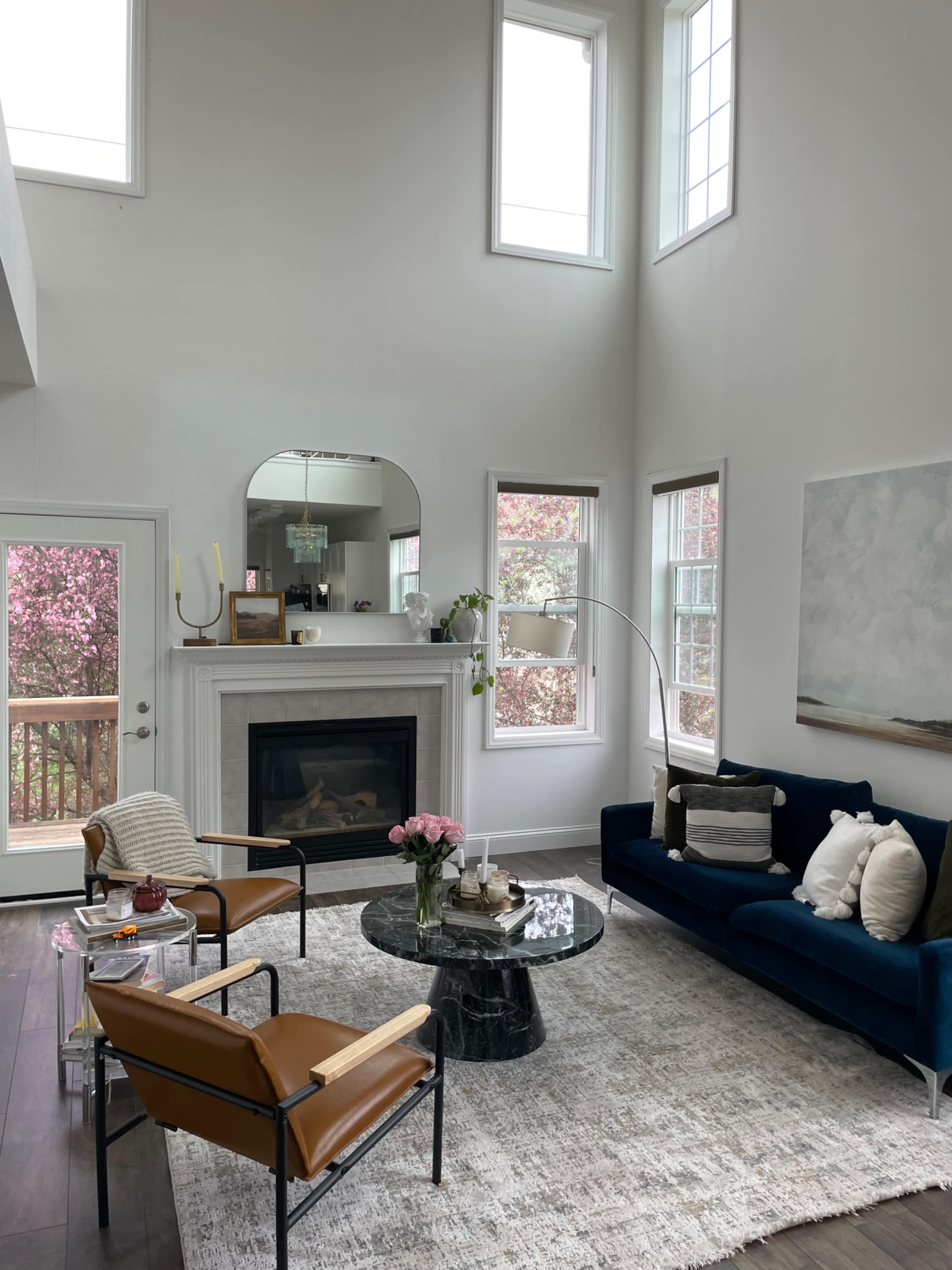 A modern living room features a marble fireplace, large windows, a navy sofa, two leather chairs, and a round coffee table on a light area rug.