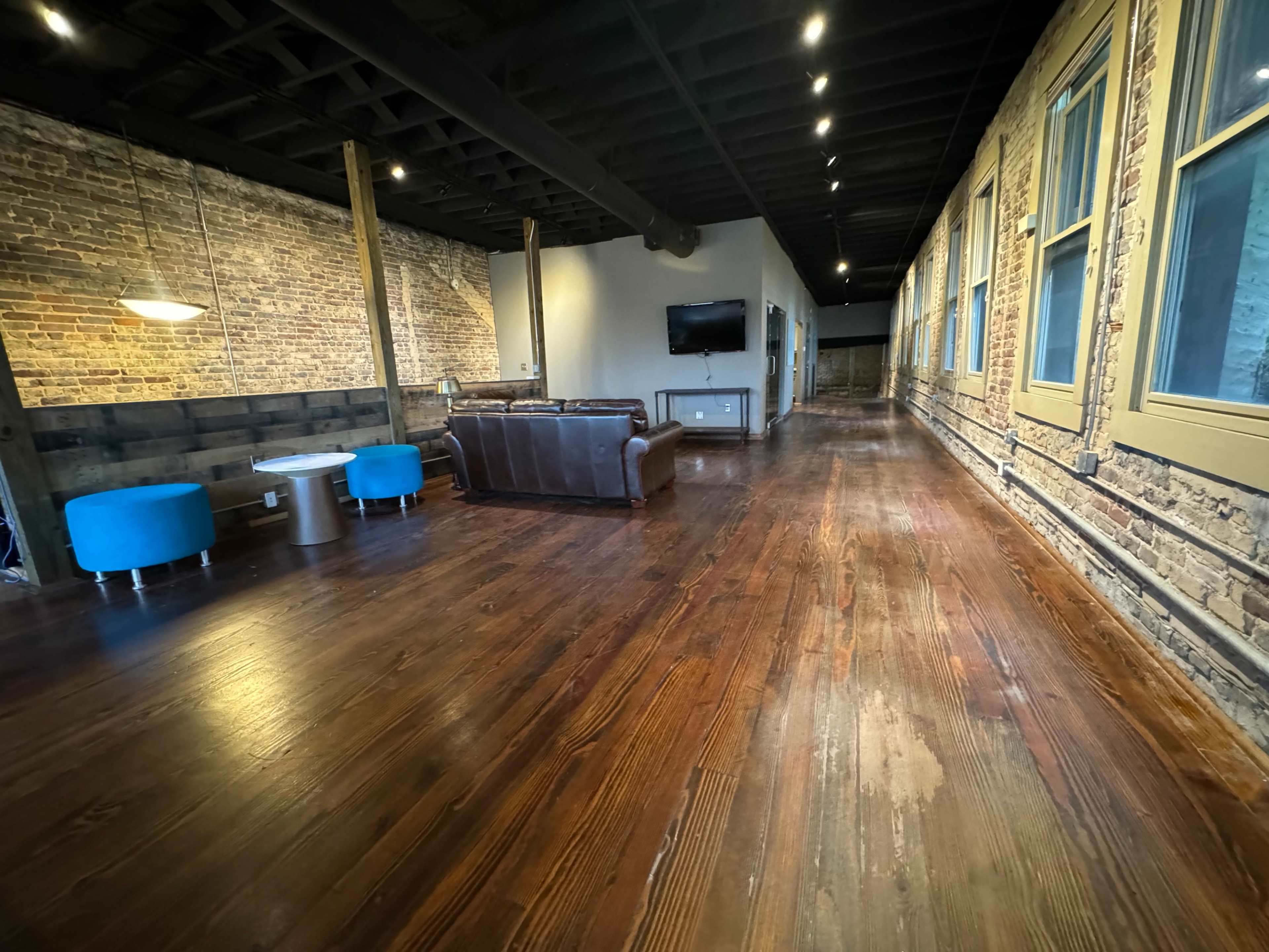 The image shows a long, narrow interior space with brick walls, wooden floors, and furniture including a dark couch and blue chairs.