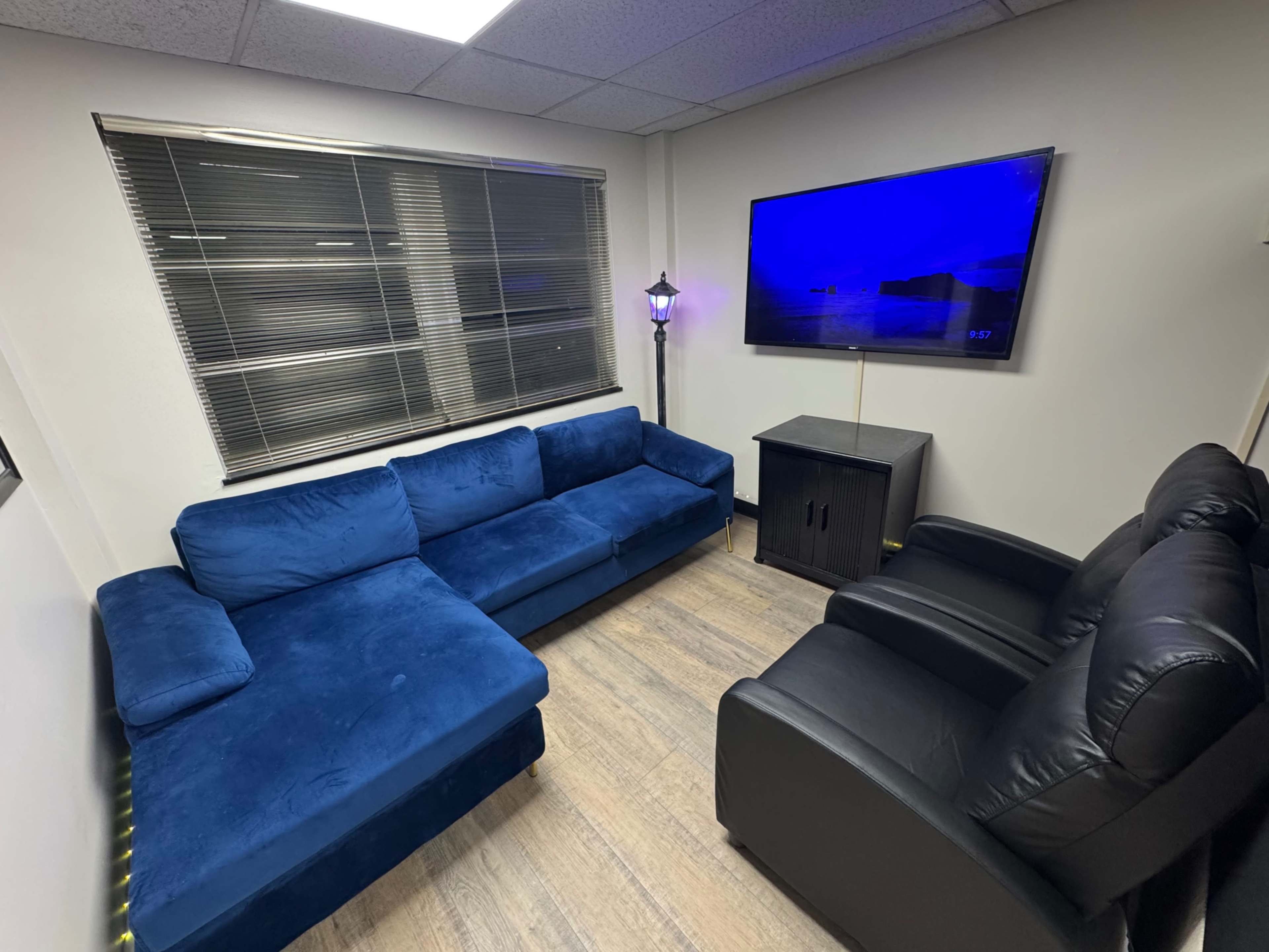 The image depicts a cozy room furnished with a blue sectional sofa, two black armchairs, a small black cabinet, and a television mounted on the wall.