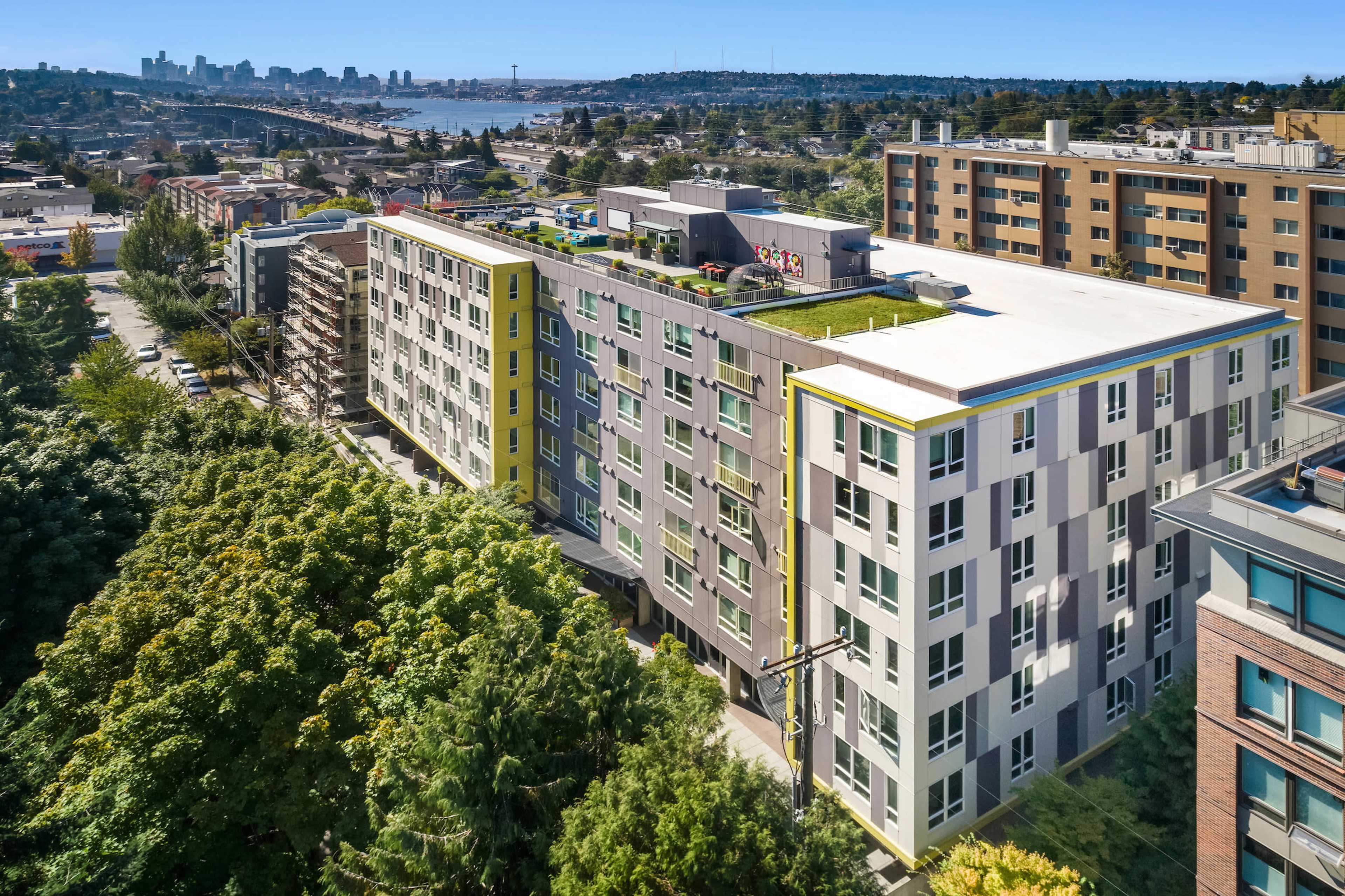 The image shows a modern, multi-story residential building with a green rooftop, situated near a city skyline and surrounded by trees.