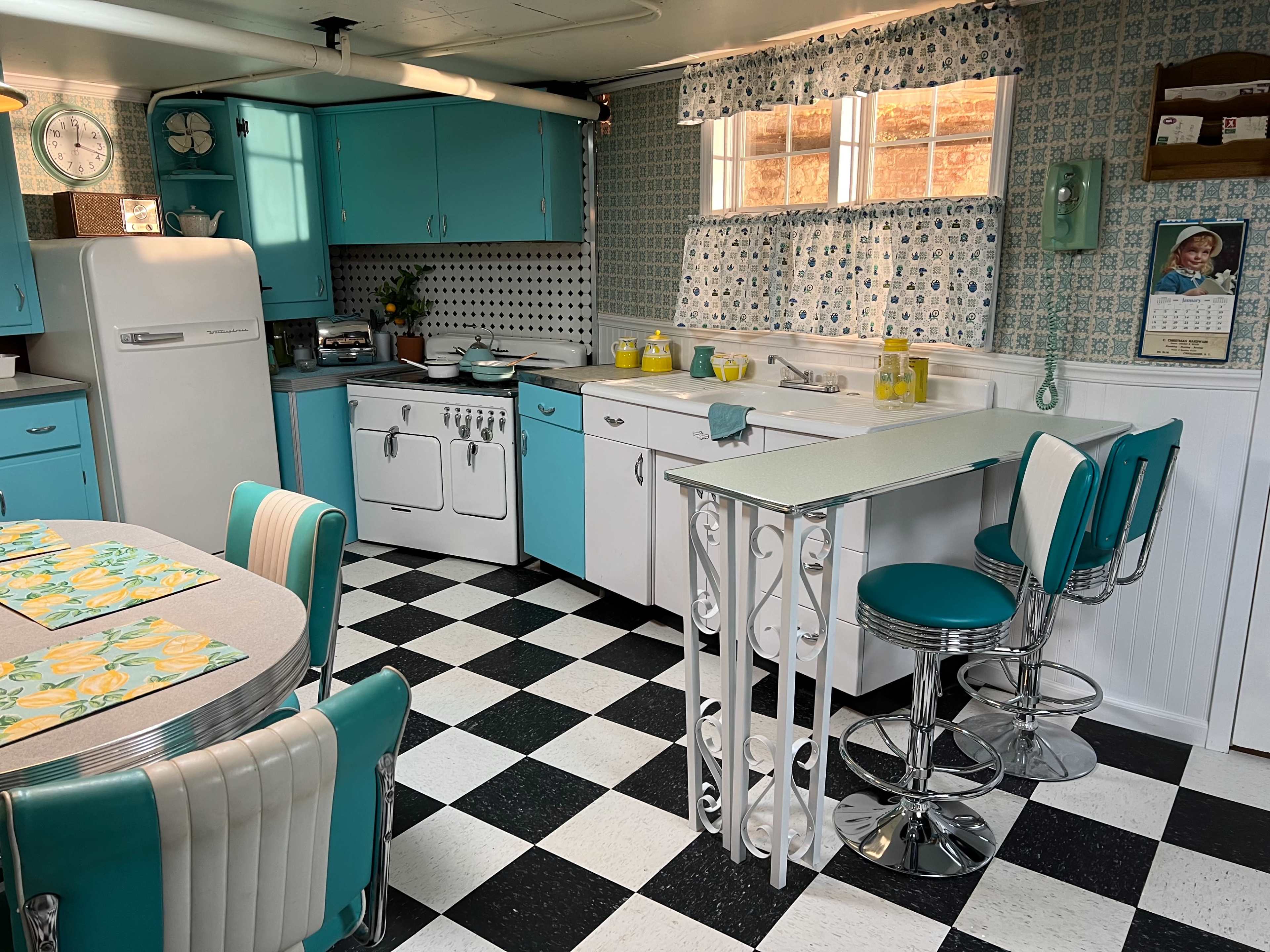 A retro-style kitchen featuring teal cabinetry, a checkered black and white floor, and a small dining table with striped chairs.