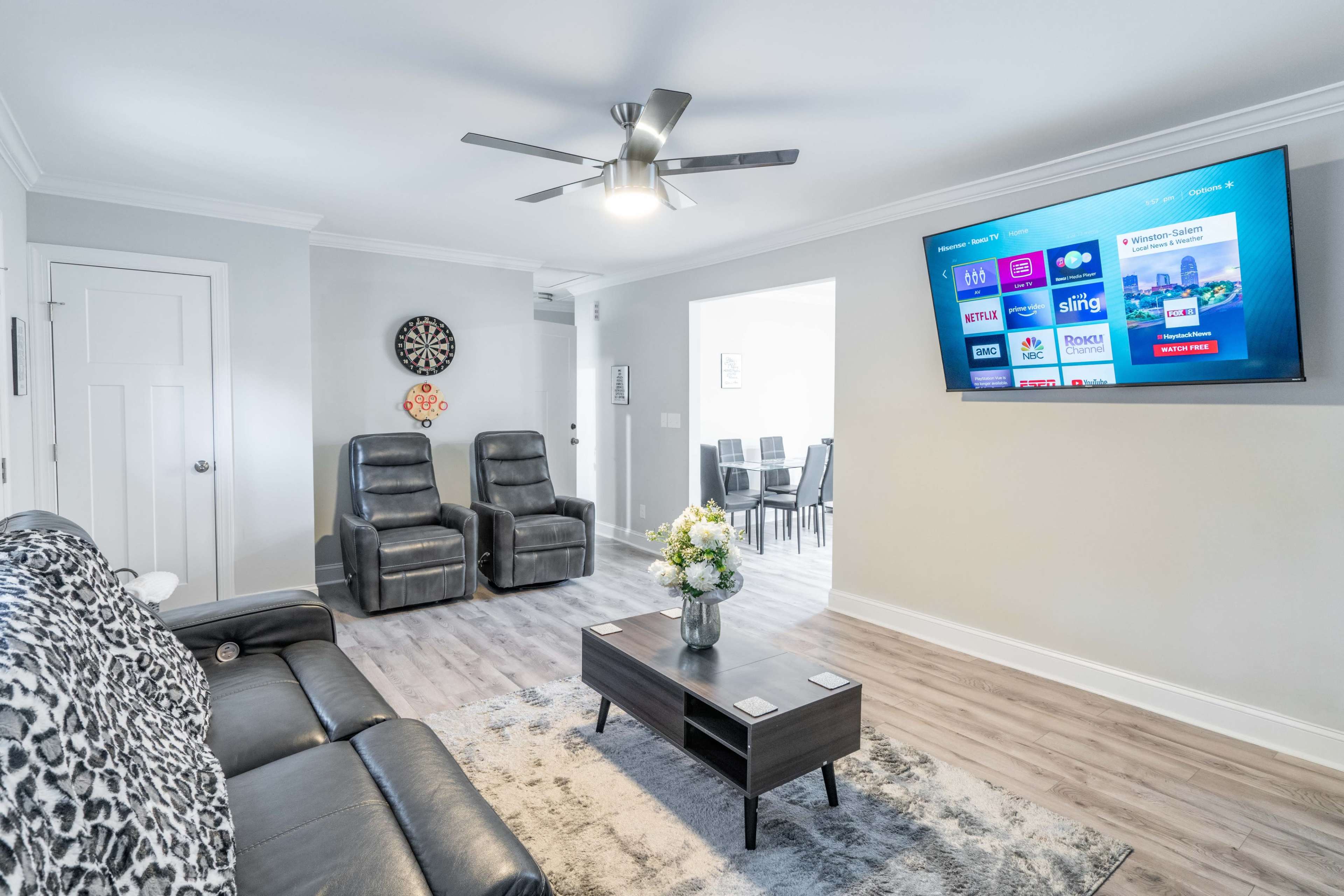 The bright living room features a large wall-mounted TV, two reclining chairs, a coffee table with flowers, and a plush area rug.