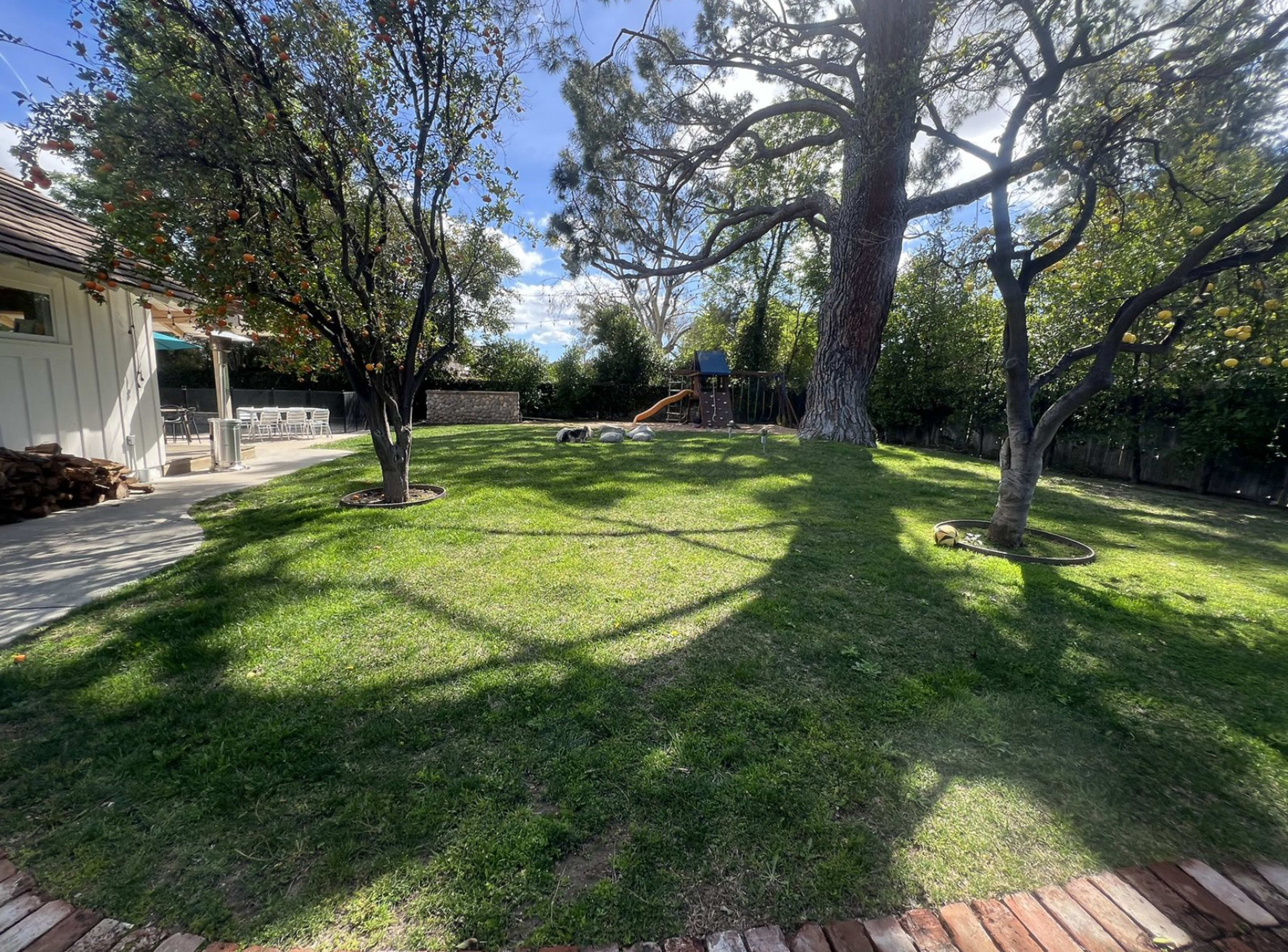 A spacious backyard features a green lawn, two trees, and a play structure visible in the background.